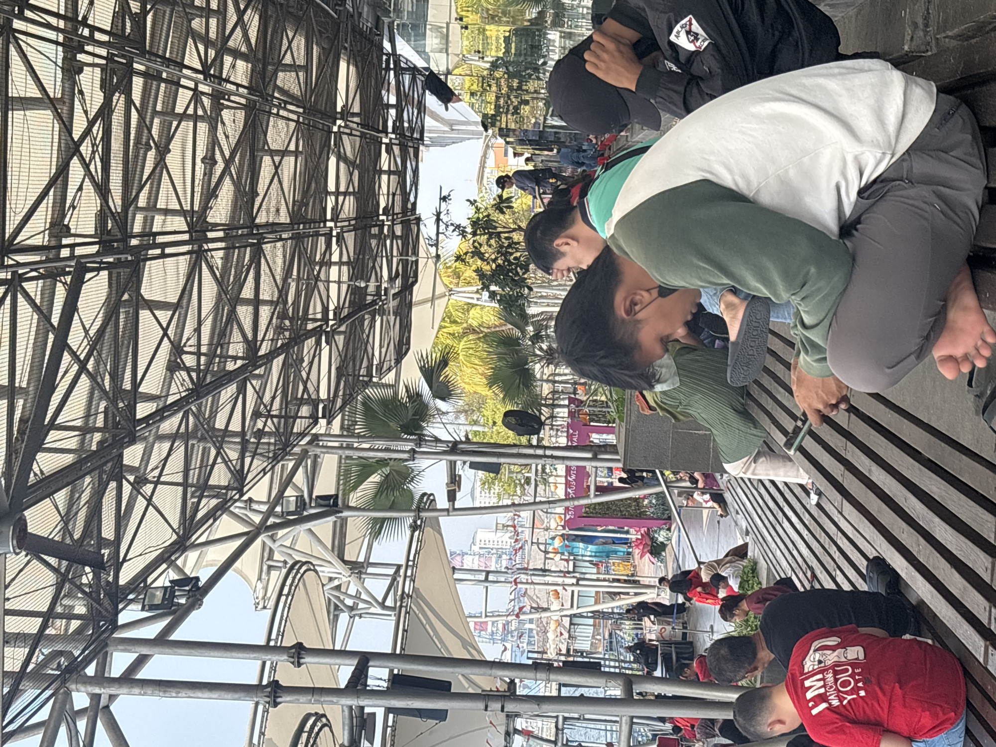 In North Jakarta, Indonesia, at an outdoor public seating area, a group of people are seated on wooden benches beneath a large, intricate metal and fabric canopy. The canopy is supported by a complex network of metal beams and poles, with translucent white fabric stretched over sections of it, allowing diffused sunlight to filter through.
In the foreground, a young man wearing a red t-shirt with the text "I'M WATCHING YOU MATE" printed on the back is sitting on a bench, facing away from the camera. To his right, a man in a black t-shirt is also seated. Further to the right, a person wearing a green and white long-sleeved shirt and grey pants is crouched on the bench, looking down at a mobile phone. Their bare feet are visible. To the far right, a person in a black jacket with patches on the sleeve is partially visible, also with their head down.
In the midground, other individuals are seated on tiered benches, some interacting with their phones, others conversing. A variety of plants, including palm trees, are visible in the background, suggesting a semi-tropical environment. Beyond the seating area, there are buildings, some with colorful facades, and a body of water with boats. The overall scene depicts a casual, public gathering space.