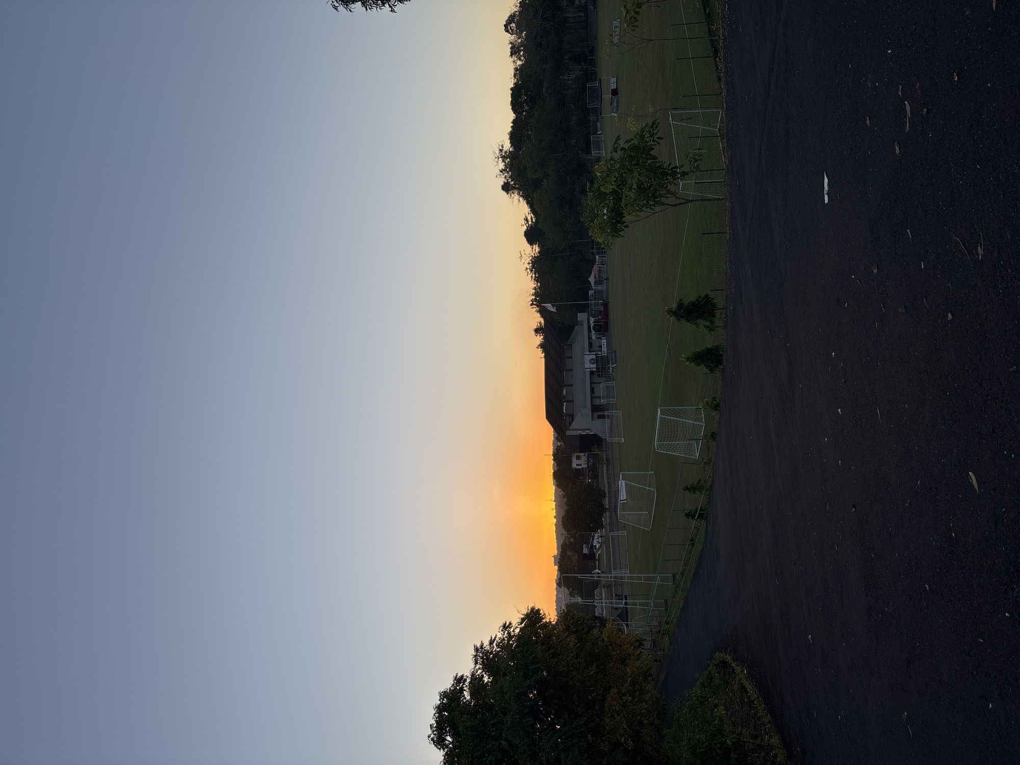 The image shows a sports field area in Kuta Selatan, Indonesia, at sunset. On the right, there is a road. In the field area, there are soccer goals visible. In the middle, trees and buildings are visible. The sky is transitioning from blue to orange, indicating dusk.
