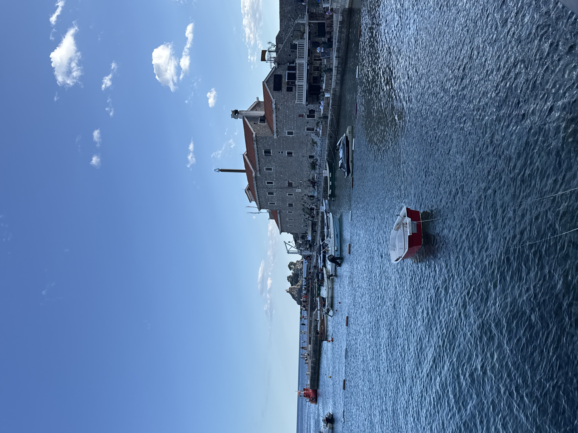 The image shows a harbor in Petrovac centar, Montenegro, on a clear day with a bright blue sky and scattered white clouds. The foreground features a calm, deep blue sea with gentle ripples. A small red and white rowboat is tethered in the middle of the water.
In the mid-ground, several other boats are moored, including smaller motorboats and a jet ski on a floating platform. A concrete pier extends into the water, lined with people. A distinctive red structure, possibly a navigational marker or a small building, is visible at the end of the pier. In the distance, a rocky island with a small structure on top is seen.
The background is dominated by a large, stone building with a red-tiled roof, likely a historic structure or hotel, situated along the waterfront. This building has multiple windows and chimneys. A terrace with outdoor seating is attached to the building, where people are seated. A stone wall runs behind the building, and a staircase leads up to it. The scene suggests a peaceful, coastal town atmosphere.