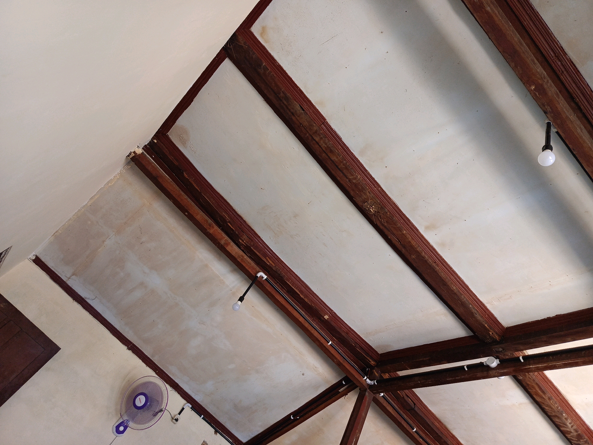 The image shows the interior ceiling of a building in Kebonagung, Indonesia. The ceiling is constructed with wooden beams, painted white panels, and exposed electrical wiring. Two bare light bulbs are visible, suspended from the wiring. A purple electric fan is mounted on a wall near the bottom left of the frame. The overall impression is of a utilitarian and somewhat rustic interior space.
