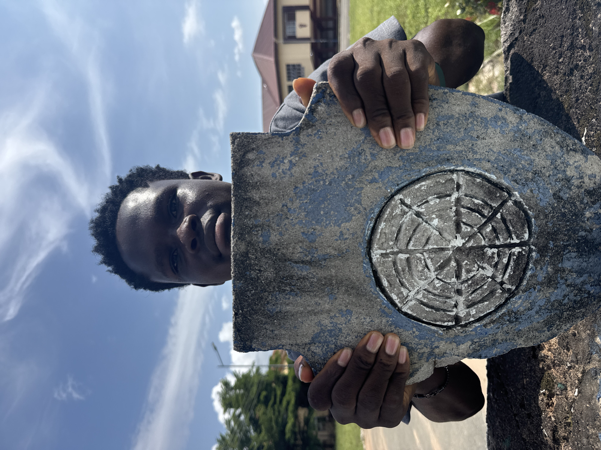 The image, oriented vertically (rotated 90 degrees clockwise), captures a close-up of a person holding a distinct object outdoors in Ihiagwa, Nigeria.
The main subject is a young, dark-skinned person, likely male, with short dark curly hair. Their face is partially visible on the left side of the frame, looking directly towards the viewer with a neutral expression. Their hands, with light pink fingernails, are prominently displayed, grasping a roughly shaped, weathered stone or concrete slab.
The object held by the person is a prominent feature. It is a thick, irregularly cut piece of material, predominantly grey with patches of faded blue paint, suggesting age and exposure to elements. On its surface, a circular design is embossed or carved in relief. This symbol resembles a stylized globe or compass rose, featuring interconnected lines radiating from a center, creating a grid-like pattern.
In the background, under a bright blue sky dotted with wispy white clouds, a multi-story building with a reddish-brown hip roof and white-framed windows is visible. Lush green grass and distant trees suggest a well-maintained outdoor environment, possibly a residential or institutional area. The lighting indicates it is daytime, likely bright and sunny. No specific text is visible on the object or in the background. The person appears to be presenting the object to the camera or using it as a prop.