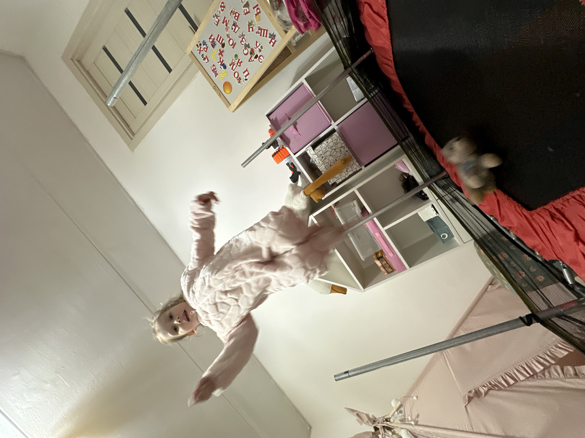 An energetic young child, likely female, is captured mid-air, possibly jumping on a trampoline in an indoor setting. She wears a light pink, textured loungewear or pajamas, and her face shows an expression of joy or concentration.
The scene appears to be a child's playroom or bedroom. A significant portion of a black trampoline mat with red safety padding is visible on the right and bottom of the frame. A small, light-colored stuffed animal rests on the trampoline mat. Behind the child and trampoline, a white shelving unit with multiple cubbies holds various items, including pink fabric storage bins and a toy gun (possibly a Nerf blaster).
On the wall above the shelving unit hangs a whiteboard adorned with numerous magnetic Cyrillic letters (e.g., M, E, Ы, Д, O, T, Ц) and numbers, along with a sun-shaped magnet, providing a strong cultural cue for the Russian location. The ceiling features a white wooden structure with dark slats, possibly an attic vent. A silver metal bar, likely part of the trampoline's safety enclosure or a room fixture, runs diagonally through the image. In the bottom right, a soft pink canopy or draped fabric is partially visible. The overall lighting suggests it is daytime or a well-lit interior. The dynamic pose of the child indicates active play.