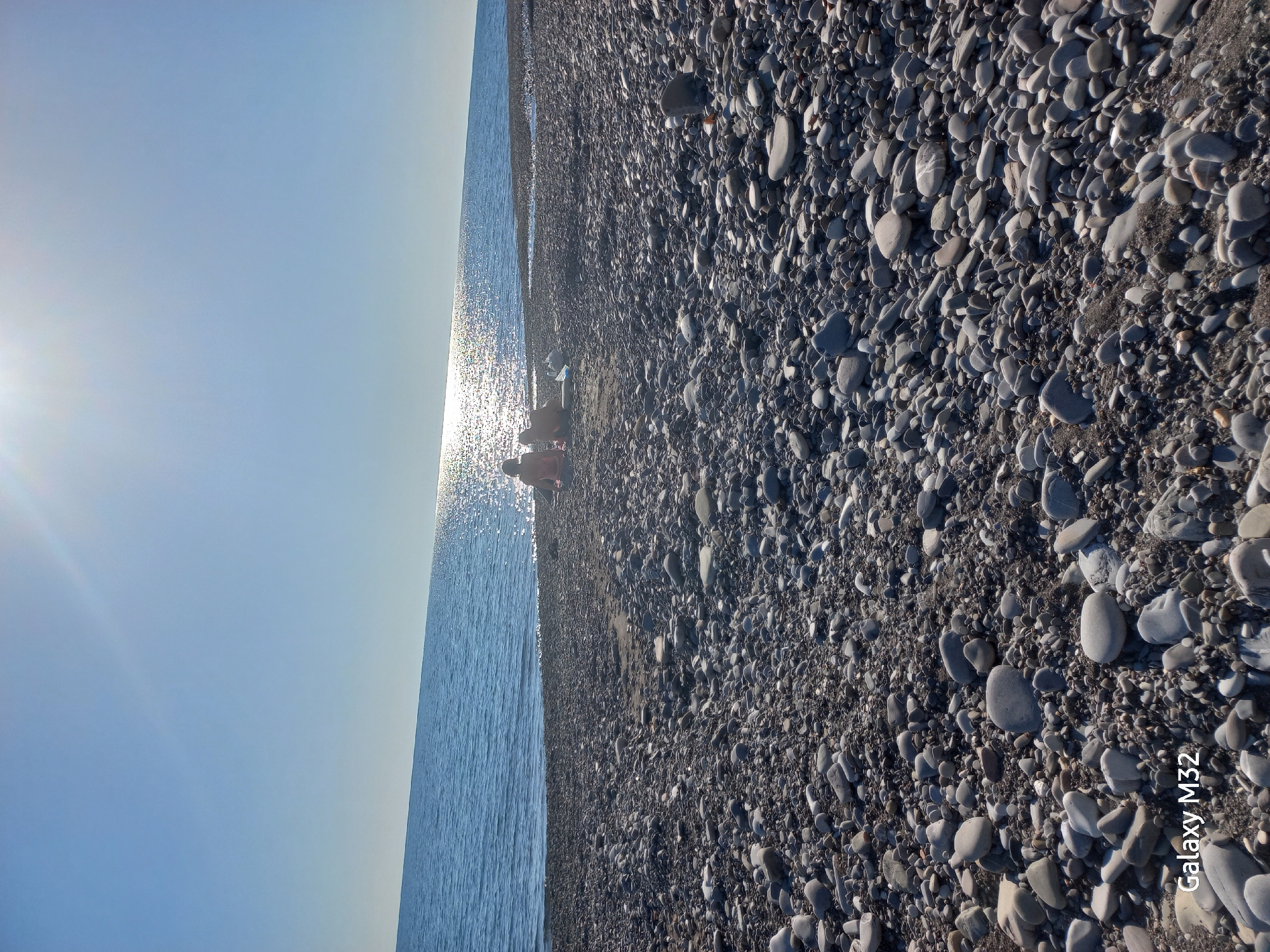 The image depicts a beach in Sochi, Russia, covered in smooth, grey and white pebbles of varying sizes. The Black Sea stretches along the left side of the image, reflecting the sunlight. Two indistinct figures, likely people, sit on the shoreline. The sky is clear and blue. A smartphone logo, "Galaxy M32", appears in the bottom right corner.