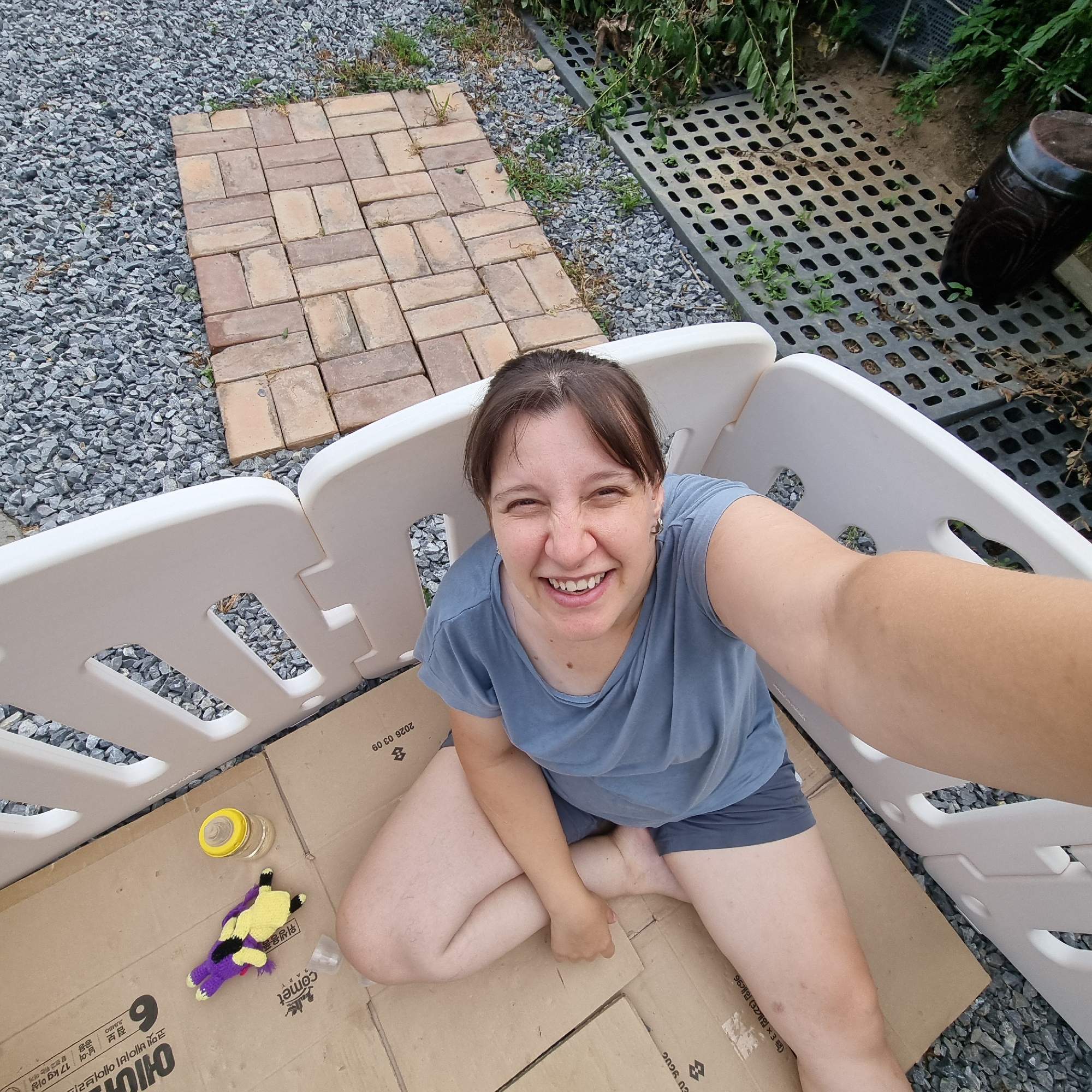In Dunnae-myeon, South Korea, a woman is positioned inside a white playpen, seated cross-legged on a cardboard floor. She is wearing a blue t-shirt and dark shorts. Her hair is pulled back, and she is smiling directly at the camera. To her left, on the cardboard, are a small yellow-capped bottle and a purple and yellow knitted item, possibly a toy. The playpen is outdoors, with gravel and a brick pathway visible in the background. A dark, large ceramic pot and a black grated platform are also present in the surrounding environment. The image appears to be a selfie, taken from a high angle.