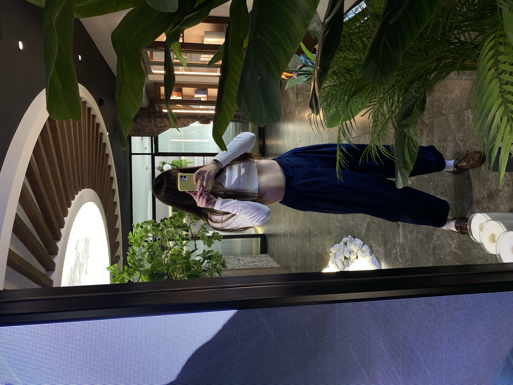 A woman with long dark hair is taking a mirror selfie in a modern indoor space with marble flooring and large plants. She is wearing a white long-sleeved crop top, dark blue wide-leg pants, white socks, and brown loafers. She is holding a yellow smartphone to take the picture. In the background, there is a large digital screen on the left, a ceiling with wooden beams, and what appears to be a retail display or shop through glass doors on the right. The lighting suggests it is daytime. The location is not identifiable as Votkinsk, Russia.