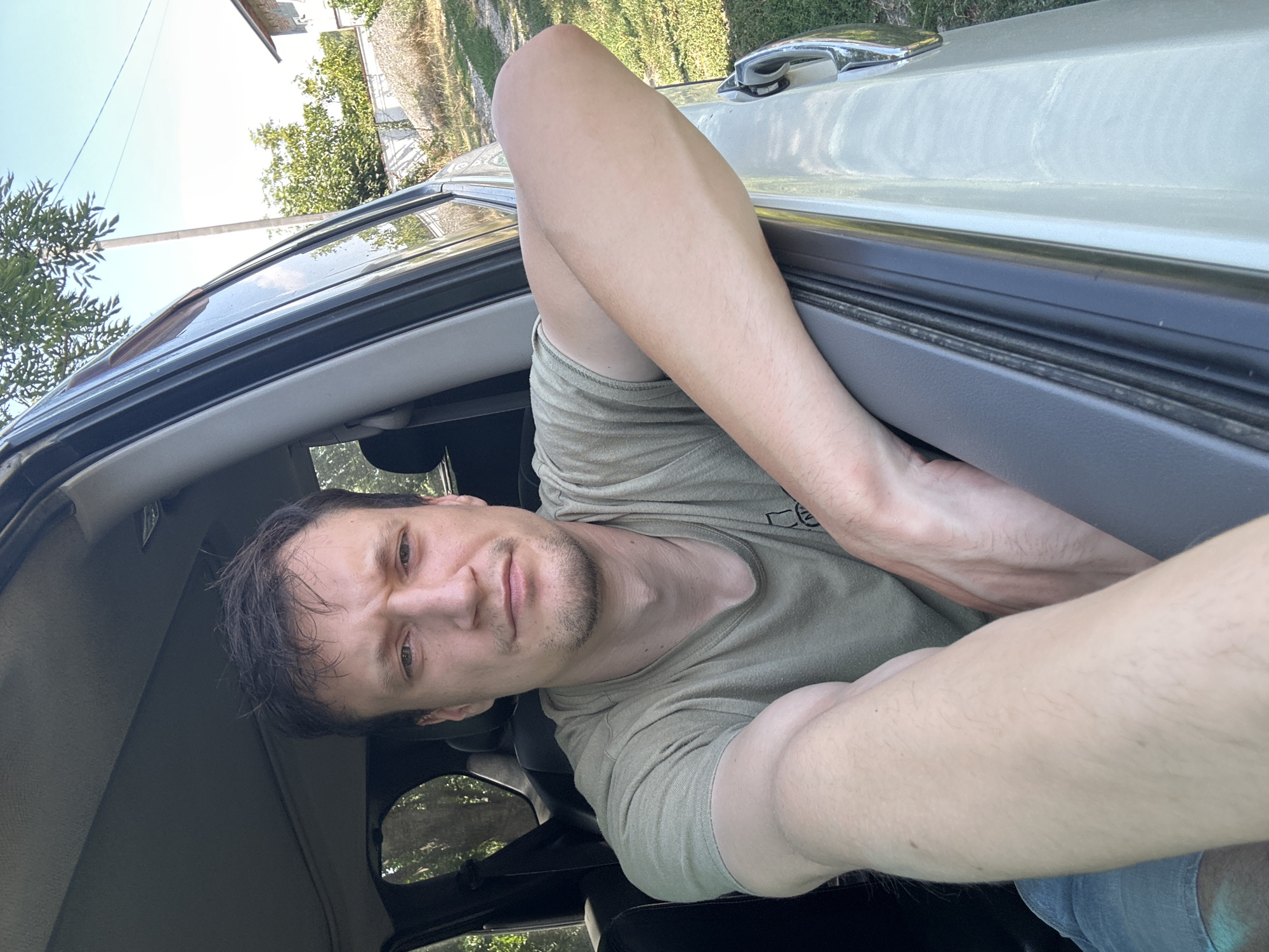 A man is sitting in the driver's seat of a car, with his head and upper body visible through the open driver's side window. He is looking towards the camera with a neutral expression. His left arm is resting on the car door frame, and his right arm is bent at the elbow, with his hand resting on his leg. He is wearing a light green t-shirt and blue denim shorts. The car is parked outdoors, with trees, a fence, and a house visible in the background. The sky is clear and blue. The image appears to have been taken in Makiv, Ukraine, based on the provided context.