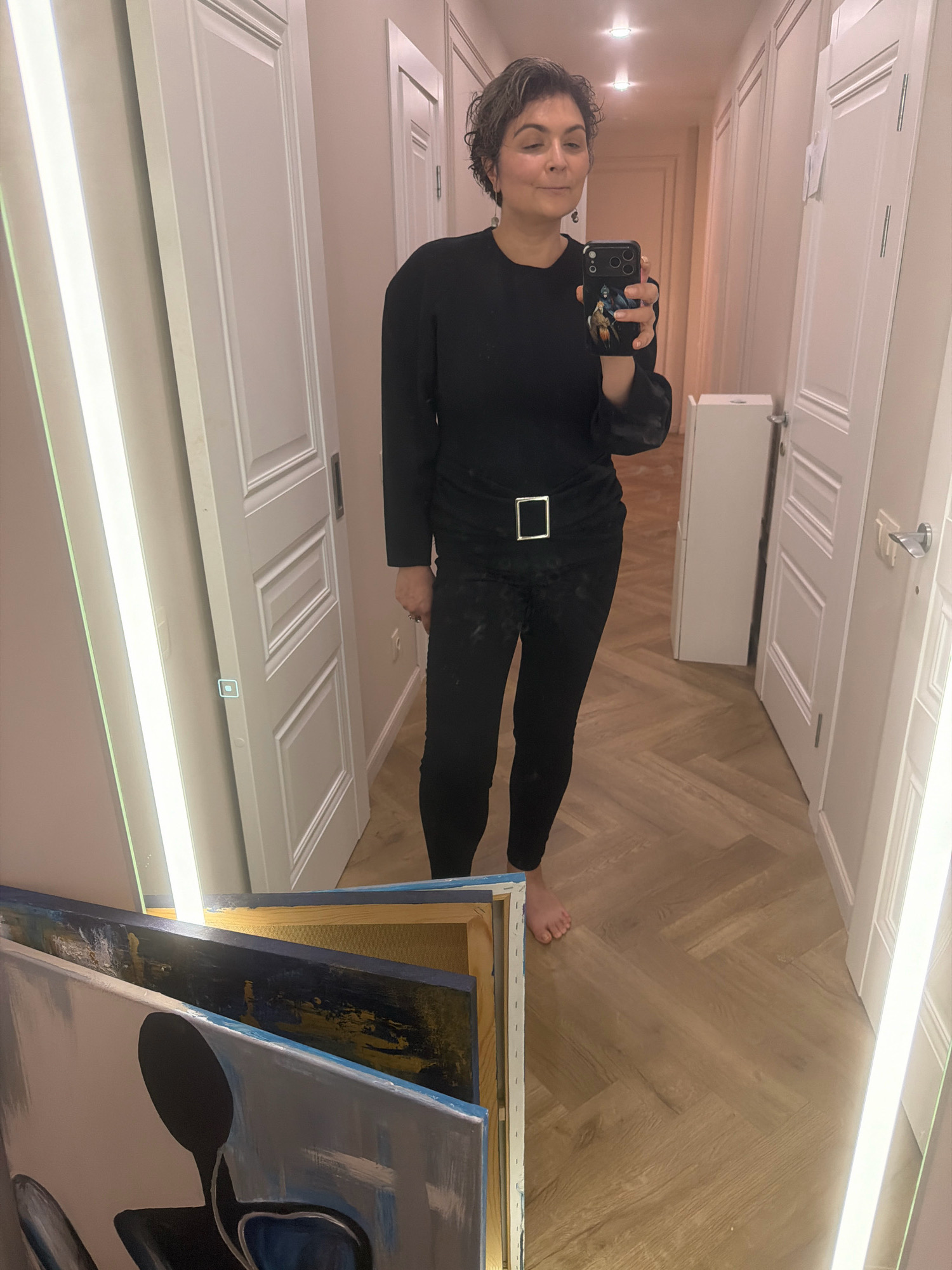The image shows a woman standing in a hallway, taking a mirror selfie. She is wearing a black long-sleeved top and black pants with a prominent square buckle belt. The hallway has light-colored walls, white paneled doors, and a herringbone pattern hardwood floor. There are tall, illuminated vertical fixtures on either side of the frame, likely part of the building's design. To the left foreground, several canvases with abstract paintings are leaning against a surface. The woman has short, curly dark hair and is holding a smartphone in her right hand. The overall impression is of a residential or commercial interior space, possibly a home or a fitting room, with an artistic element present in the foreground. This image does not contain any identifiable landmarks or features that would allow for a specific location in Samara, Russia, to be determined.