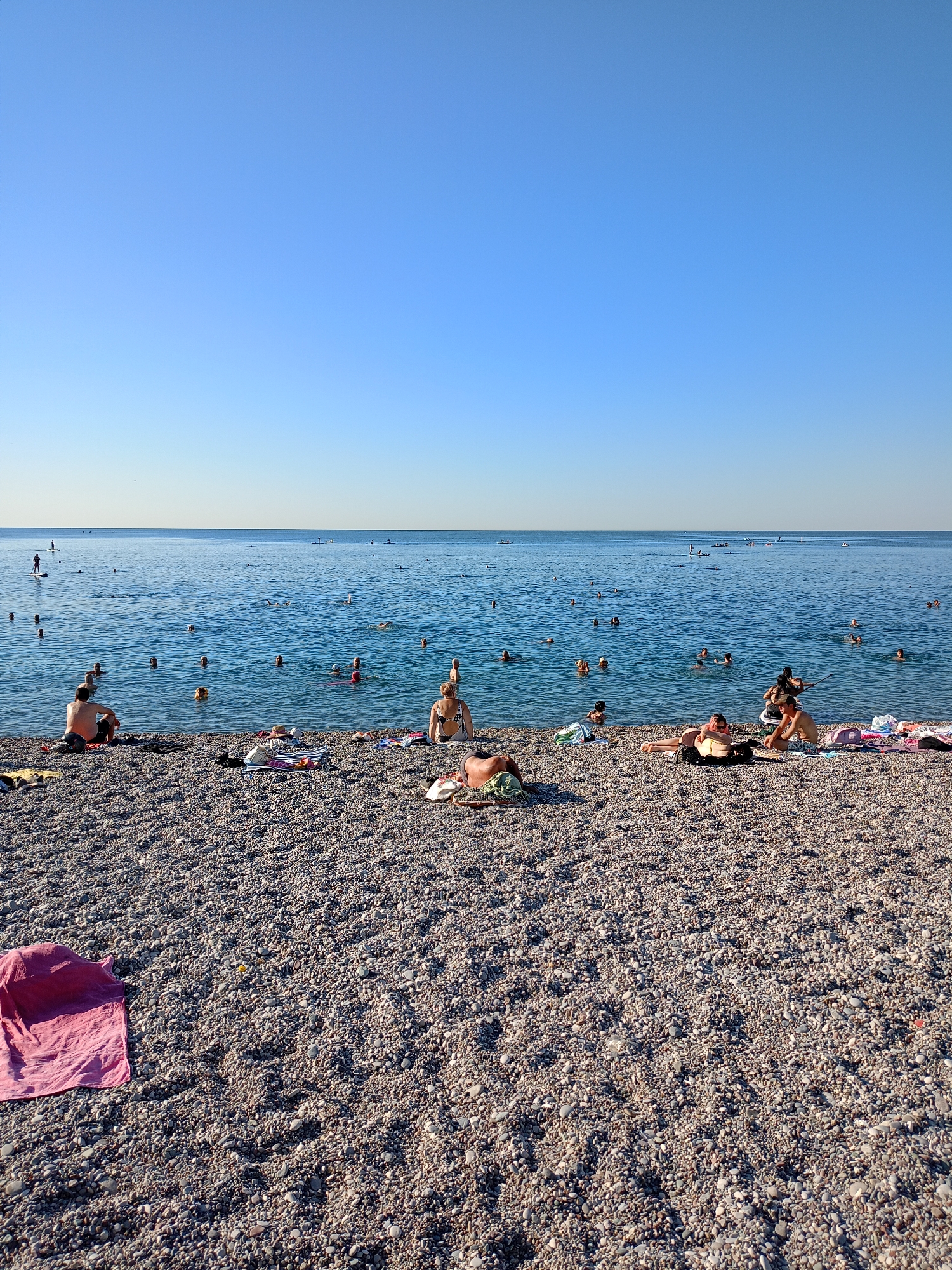 This is a full-shot photograph of a crowded pebble beach on the coast of Antalya, Turkey, under a clear blue sky. The water is calm and blue, with many people swimming and wading. Several individuals are sunbathing on the pebbles, some with towels and bags. In the distance, there are people on paddleboards. The scene appears to be during daylight hours.