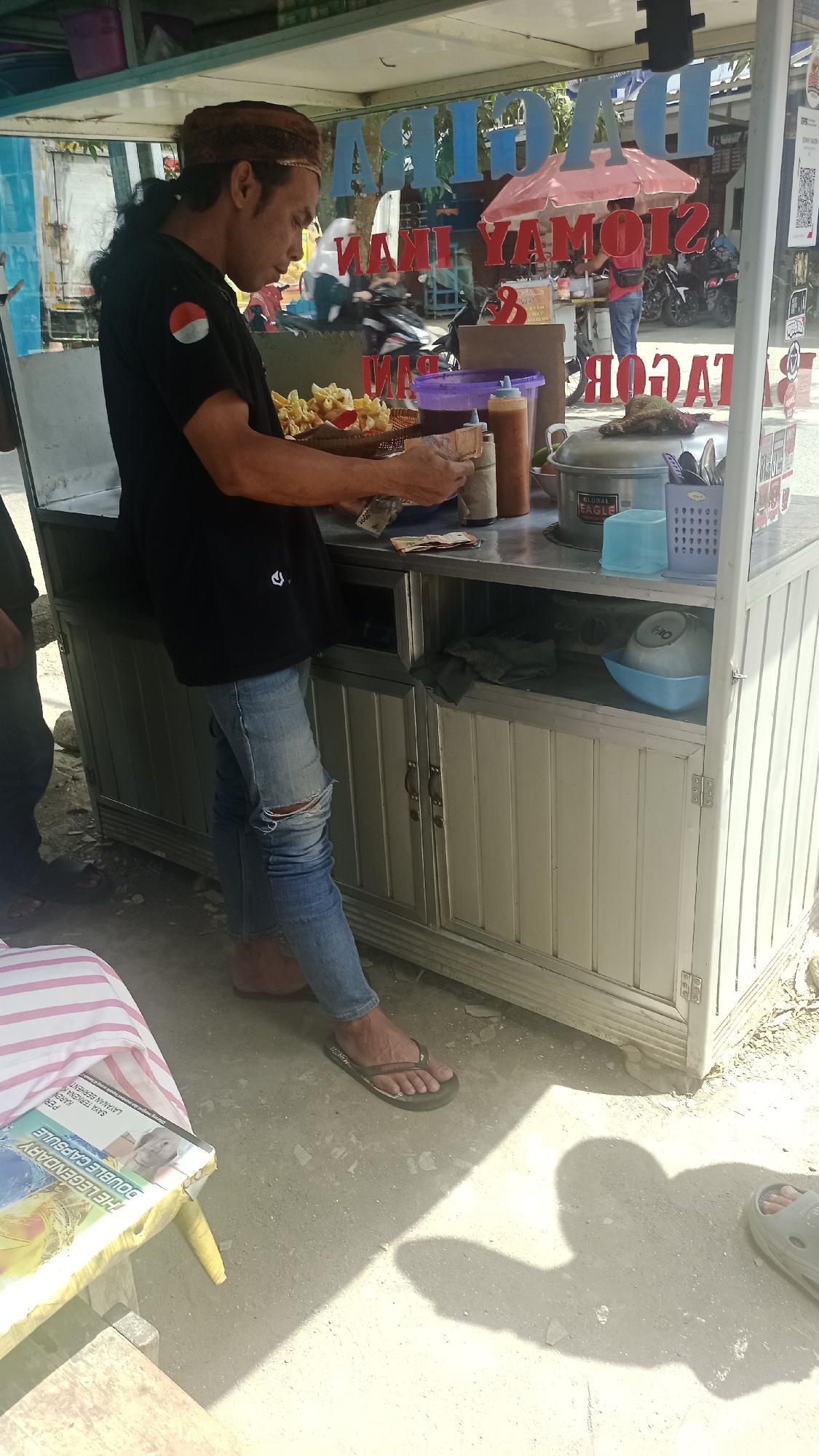 A man stands behind a food cart in Cibeber, Indonesia. The cart is white and silver, with glass panels on the front displaying colorful text and images. The man, wearing a black t-shirt with an Indonesian flag emblem, ripped blue jeans, and a brown head covering, is handling money and food items. In front of him on the cart are various condiments, a basket of fried food, and kitchen utensils. Behind him, the street is visible with other food vendors and vehicles. The scene suggests a bustling street food environment.