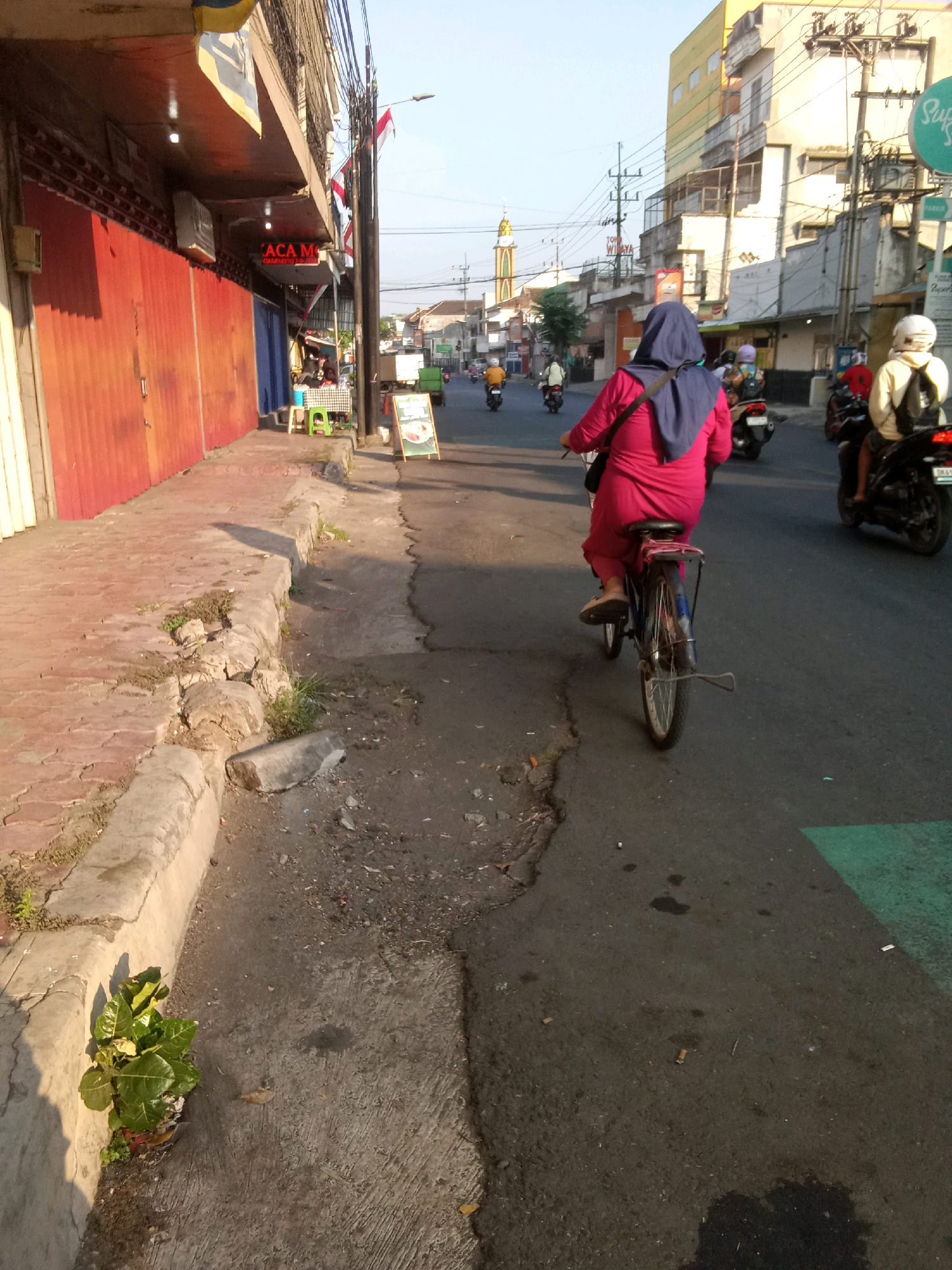 A woman in a bright pink dress and a blue hijab rides a bicycle along a busy street in Jember, Indonesia. She is on the right side of the road, facing away from the camera. The street is lined with buildings on the left, some of which have red metal shutters. There are other vehicles and people on the street, including motorcycles. The ground is paved with asphalt and has some debris scattered on it. There is a sidewalk on the left side of the street, made of interlocking concrete pavers and a concrete curb. The sky is clear and blue.