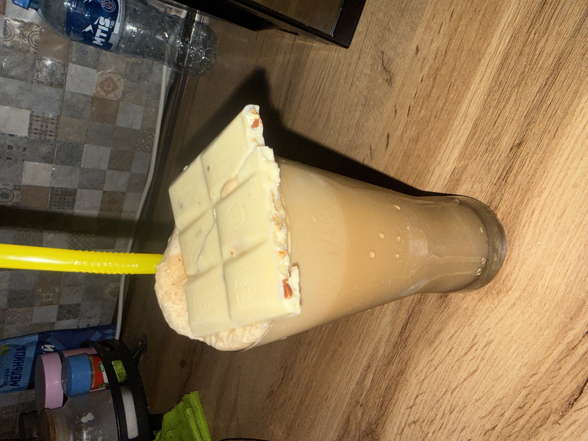 This media file shows a close-up, angled shot of a tall glass filled with a light brown, frothy beverage. A large piece of white chocolate, broken into sections and appearing to have almonds embedded within it, rests on top of the drink, partially submerged. A bright yellow plastic straw is inserted into the drink, extending upwards.
The setting appears to be indoors, with a wooden surface in the foreground, likely a table, exhibiting a light wood grain pattern. In the background, a tiled surface is visible, with a pattern of small, square tiles in various shades of grey, brown, and cream. A partially visible plastic bottle with the label "MTIS" and some blue liquid is also in the background, positioned behind the tiled area. Other small objects, including what appear to be containers or jars with colored lids, are present in the lower left portion of the frame.
The lighting suggests an indoor environment, possibly with artificial light. The overall mood is casual, with the focus on the decadent drink. No people are visible in the frame. The visible text includes "MTIS" on the bottle and a partial label in Cyrillic script on a blue package in the background, which appears to read "МЕЛЬНИЦА". The location context of Tbilisi, Georgia is provided.