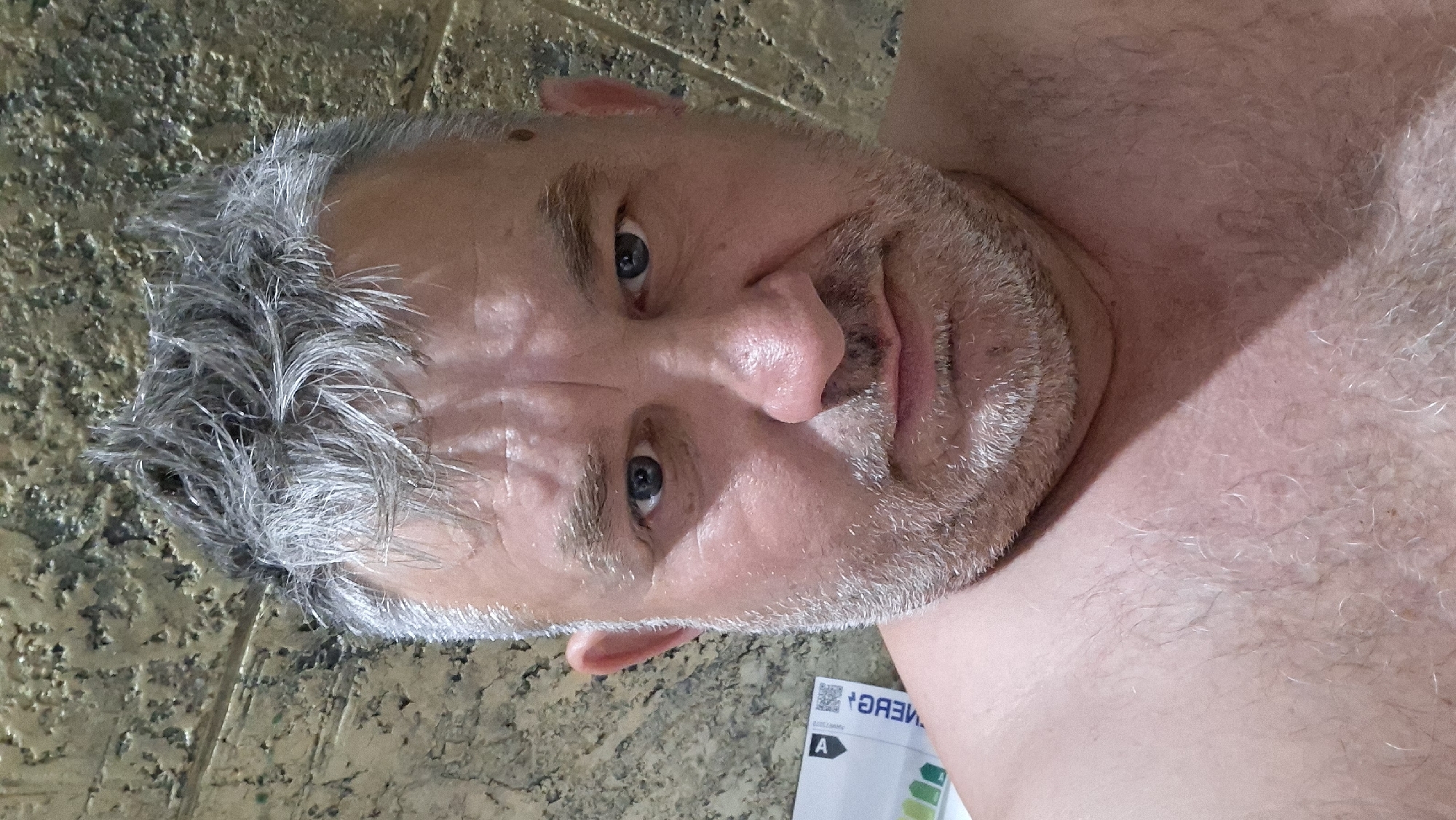 A close-up, selfie-style photograph shows a man from the chest up. He has short, messy gray hair, prominent wrinkles on his forehead, and a short, gray beard. His eyes are blue, and he has a serious expression. He is shirtless, revealing light chest hair. The background is a textured wall in a warm, earthy tone. A small portion of a white label with black text and colored bars is visible on the left side of the image. The lighting suggests an indoor setting. There are no visible signs of injury or distress. The scene does not contain any specific elements that would allow for its precise location to be determined within Voinova, Moldova.