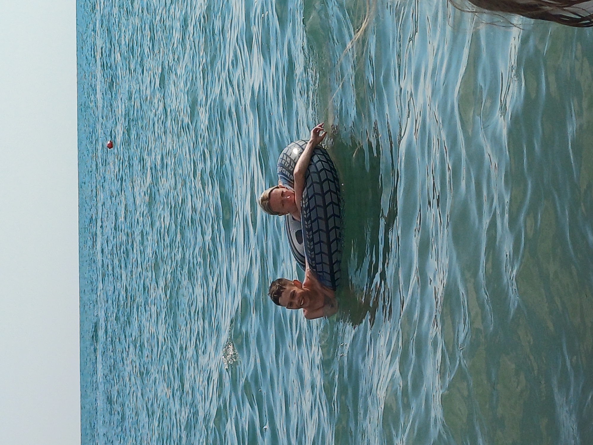The image shows two individuals, a young boy and an adult, in the Black Sea off the coast of Sochi, Russia. The boy is on the left, smiling and partially submerged in the water. The adult is on the right, leaning on an inflatable tube that is also used by the boy. The water is a deep blue with visible ripples, and the sky is a clear, pale blue. There is a small orange buoy in the distance. The scene suggests a recreational activity at sea.