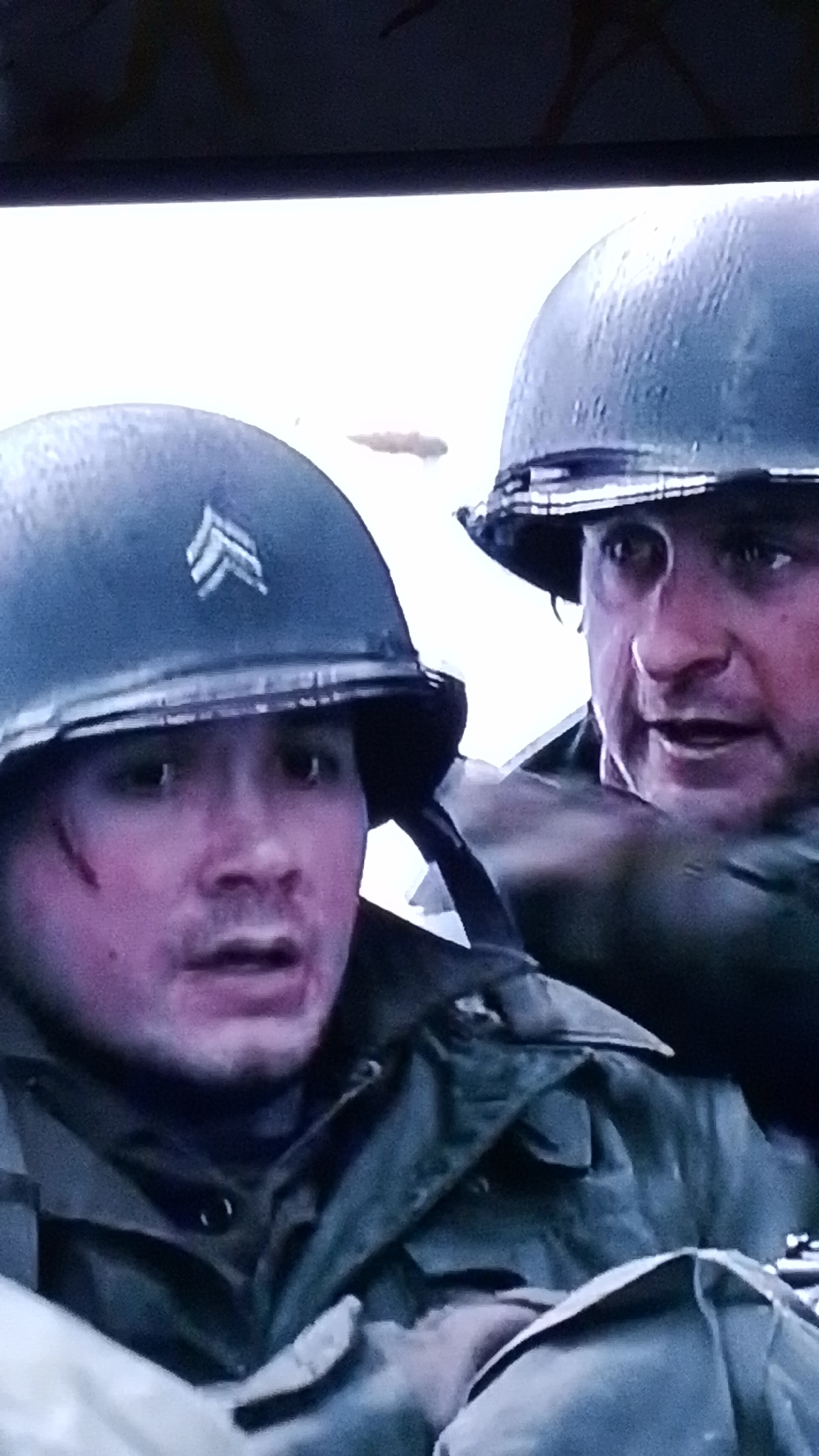 Two soldiers wearing helmets are depicted in a close-up shot. The soldier in the foreground has a wound on his face and a chevron insignia on his helmet. The soldier behind him is partially visible and appears to be speaking. The scene appears to be set in a wartime environment. There is no indication that this image relates to Tinaquillo, Venezuela.