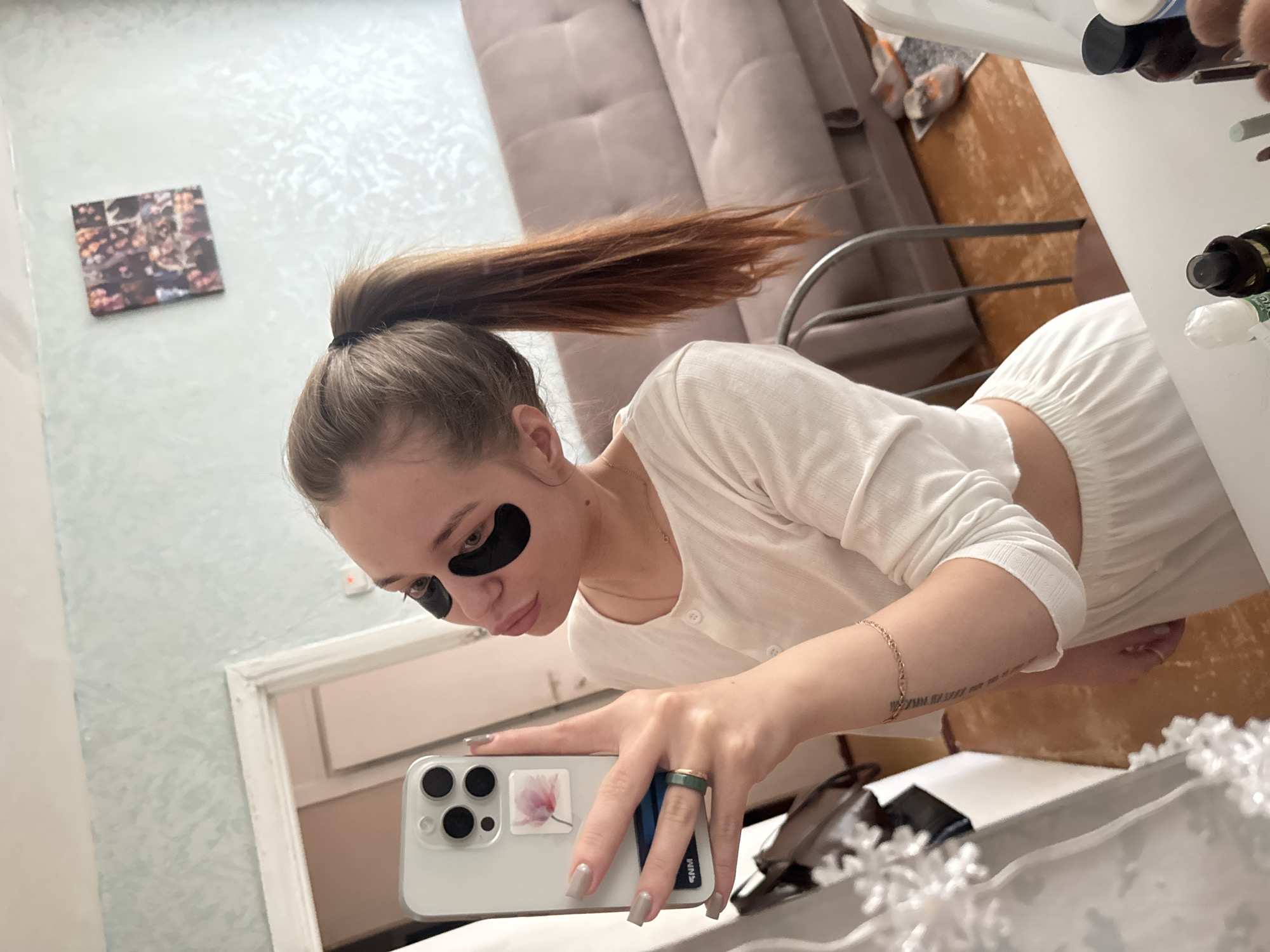 A woman is taking a selfie in a bathroom. She is wearing black eye masks and a white outfit. She has her hair pulled back in a ponytail. She is holding a white smartphone with a floral design on the back. The background includes a bathroom mirror, a cabinet, and what appears to be a portion of a couch in an adjacent room. The location may be Chaikovsky, Russia, though there is no definitive indication of this in the image.