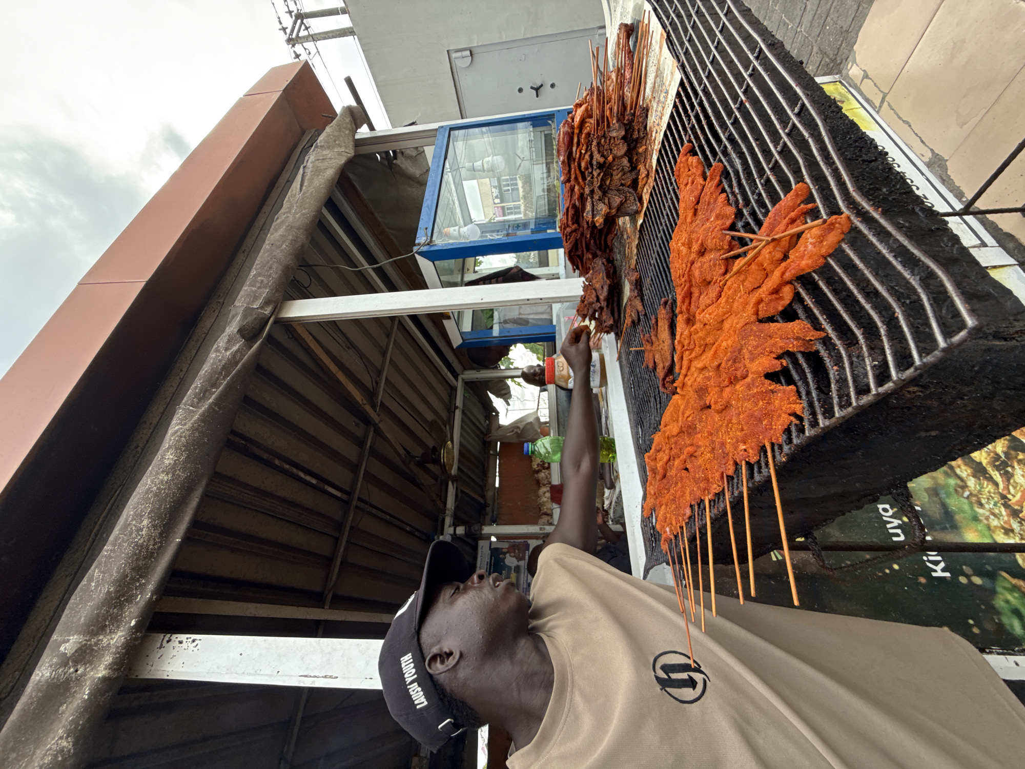 A street food vendor in Ado, Nigeria, is captured at work in this image, preparing skewers on a large grill. The man, with dark skin, wears a beige t-shirt featuring a black circular logo and a black baseball cap with "LAUSU YOUTH" written in white. He stands to the left of the grill, looking upwards with a focused, neutral expression, and his right hand is extended towards a cluster of skewers.
The grill, robust and blackened from use, is laden with numerous meat skewers. On the right side of the grill, a prominent display of bright reddish-orange skewers, likely spiced chicken or beef suya, are arranged. To their left, a pile of darker, reddish-brown skewers, possibly beef or organ meats, appear more cooked.
The setting suggests an informal outdoor stall. Behind the man and the grill, a simple structure made of corrugated metal sheets forms part of the vendor's setup. A small glass display case with a blue frame is visible above, reflecting elements of the surroundings. The sky above is overcast, indicating a cloudy day. Below the grill, a poster or sign is partially visible, showing text that includes "Ki uya," strongly suggesting an advertisement for 'suya,' a popular grilled meat dish. A partial phone number, "02 394 88," is also discernible. The scene conveys a typical bustling street food environment.