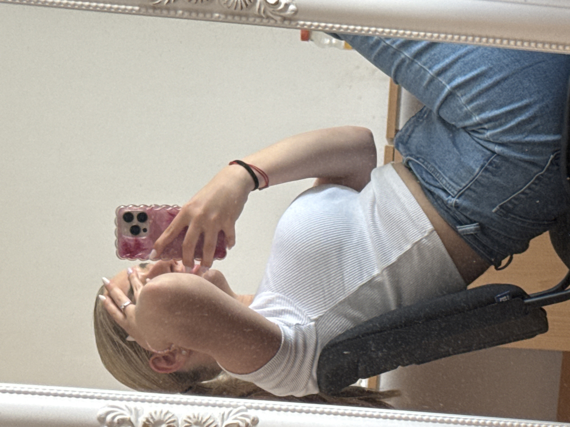 The image is a mirror selfie, rotated 90 degrees clockwise, depicting a person's reflection in an ornate white-framed mirror. The subject, a female with blonde or light brown hair, is shown upside down relative to the frame. She is wearing a white ribbed crop top and blue jeans, and is seated on a dark office chair, the backrest of which is visible behind her.
She holds a pink smartphone, encased in a ruffled clear cover, with her right hand, positioning it to capture her reflection. Her left hand, with long, white-painted nails and a silver ring on the ring finger, is near her face. A black and a red thin bracelet are visible on her left wrist.
The background reflected in the mirror is a plain, light-colored wall. A small, indistinct blue label or tag is visible on the backrest of the chair. The scene takes place indoors, likely within a room in Sofia, Bulgaria, as the person poses for a casual mirror photograph.