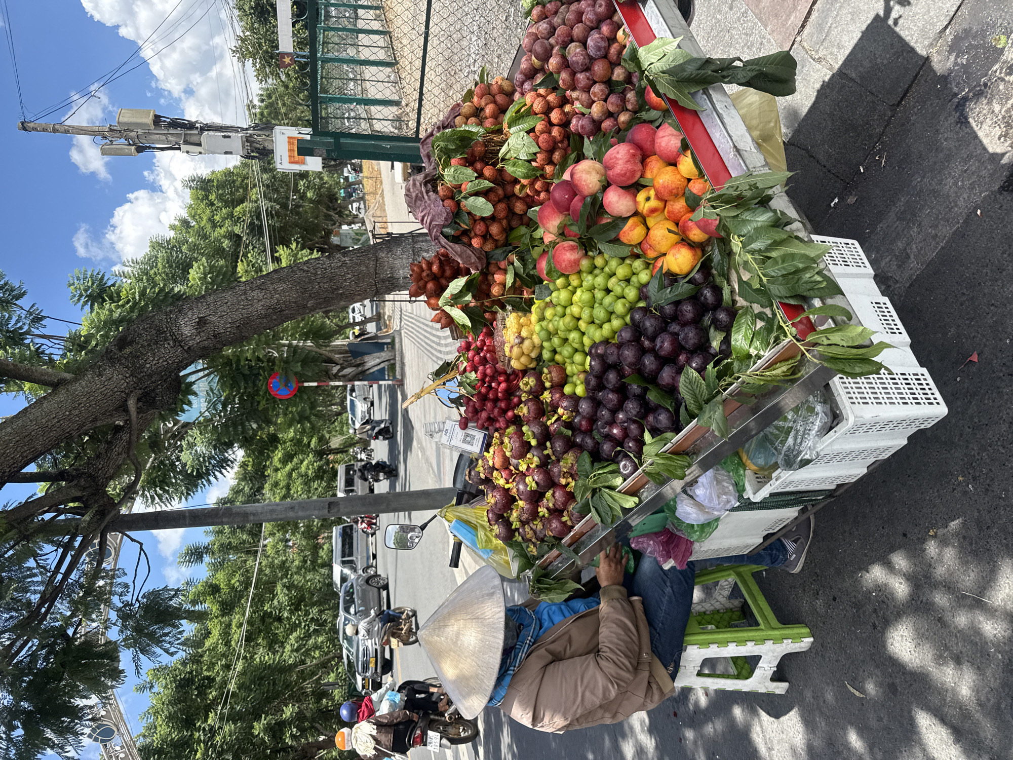 A fruit cart vendor sits on a plastic stool beside a cart laden with a variety of fruits on a street in Da Lat, Vietnam. The vendor, wearing a conical hat and a light brown jacket, is partially obscured by the cart. The cart displays an abundance of mangosteens, red and green grapes, lychees, salak, peaches, and other fruits. In the background, traffic, including motorcycles and cars, moves along the street. Trees line the sidewalk, and a cellular tower is visible in the distance. The scene is illuminated by natural sunlight, casting shadows on the ground.
