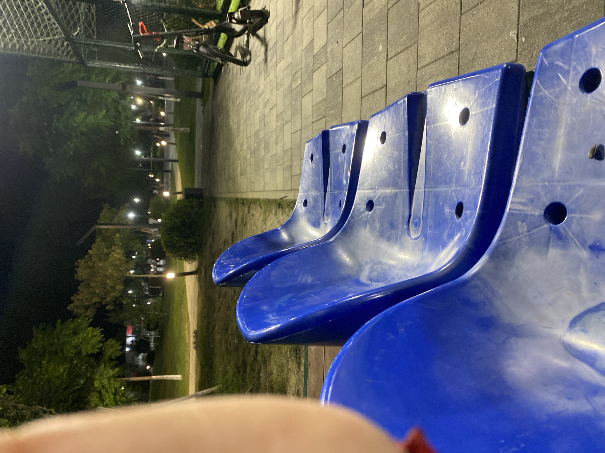 The image depicts a nighttime scene at a park or recreational area in Lagodekhi, Georgia. In the foreground, several bright blue plastic stadium seats are visible. The seats are arranged in a row and appear to be slightly worn, with some scuff marks. To the right of the seats, a paved walkway with a checkerboard pattern of grey tiles is visible, leading towards the background. Beyond the walkway, there is a fenced area, possibly a sports court, with a bicycle or scooter leaning against it. The background features trees, grass, and lampposts illuminating the area. Several indistinct figures of people are scattered throughout the park. The overall atmosphere is one of a public space at night, with a focus on the seating and paved areas.