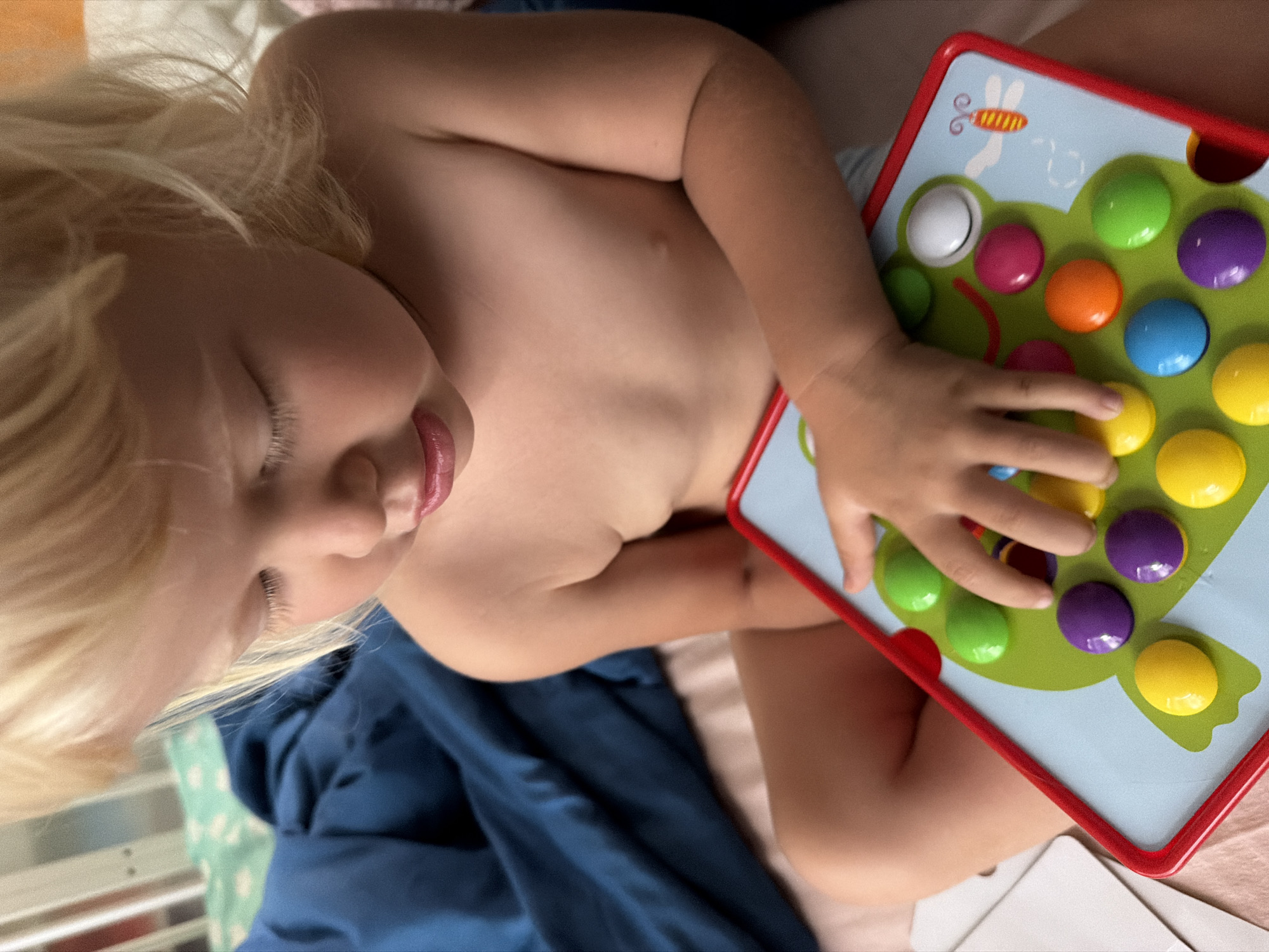 A close-up, eye-level shot shows a young Caucasian child with blonde hair and closed eyes, focused on a colorful toy frog board with large, round, multi-colored buttons, in Sochi, Russia. The child is shirtless and sits on a bed with blue and white bedding. The child's left hand is on the toy board, interacting with the buttons. The background includes a white crib and blue fabric. The lighting is soft, suggesting an indoor setting.