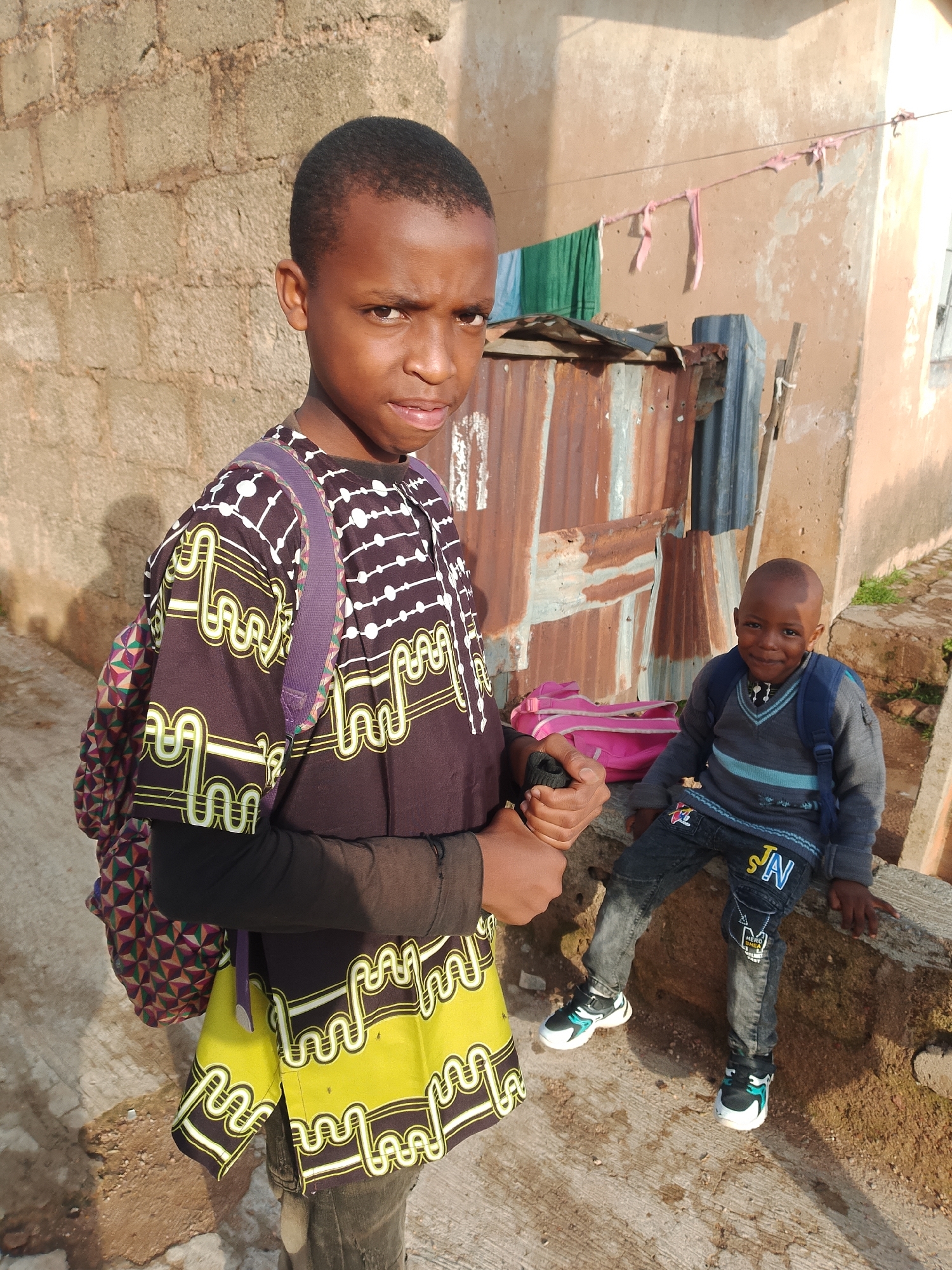 In Jos, Nigeria, an outdoor scene unfolds with two young boys near a rustic dwelling. The boy in the foreground, facing the camera, wears a dark long-sleeved shirt with a striking yellow and white patterned tunic over it, and a purple backpack. His expression is serious as he holds a black object. Behind him, a younger boy is seated on a concrete ledge, wearing a grey sweater with blue trim, dark jeans adorned with patches, and a blue backpack. A pink backpack rests beside him. The background features a building constructed with rough textured walls, corrugated metal sheeting, and laundry hanging on a wire, suggesting a lived-in environment. The lighting indicates it is daytime, with sunlight casting shadows.