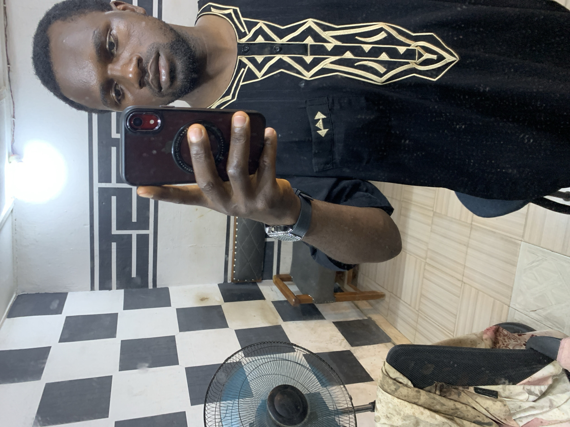 A man with dark skin and short black hair is taking a selfie in a room that appears to be in Jos, Nigeria. He is wearing a black garment with gold embroidery. In the background, there is a checkered pattern on the wall, a fan, and a chair. The room is lit by an overhead light.