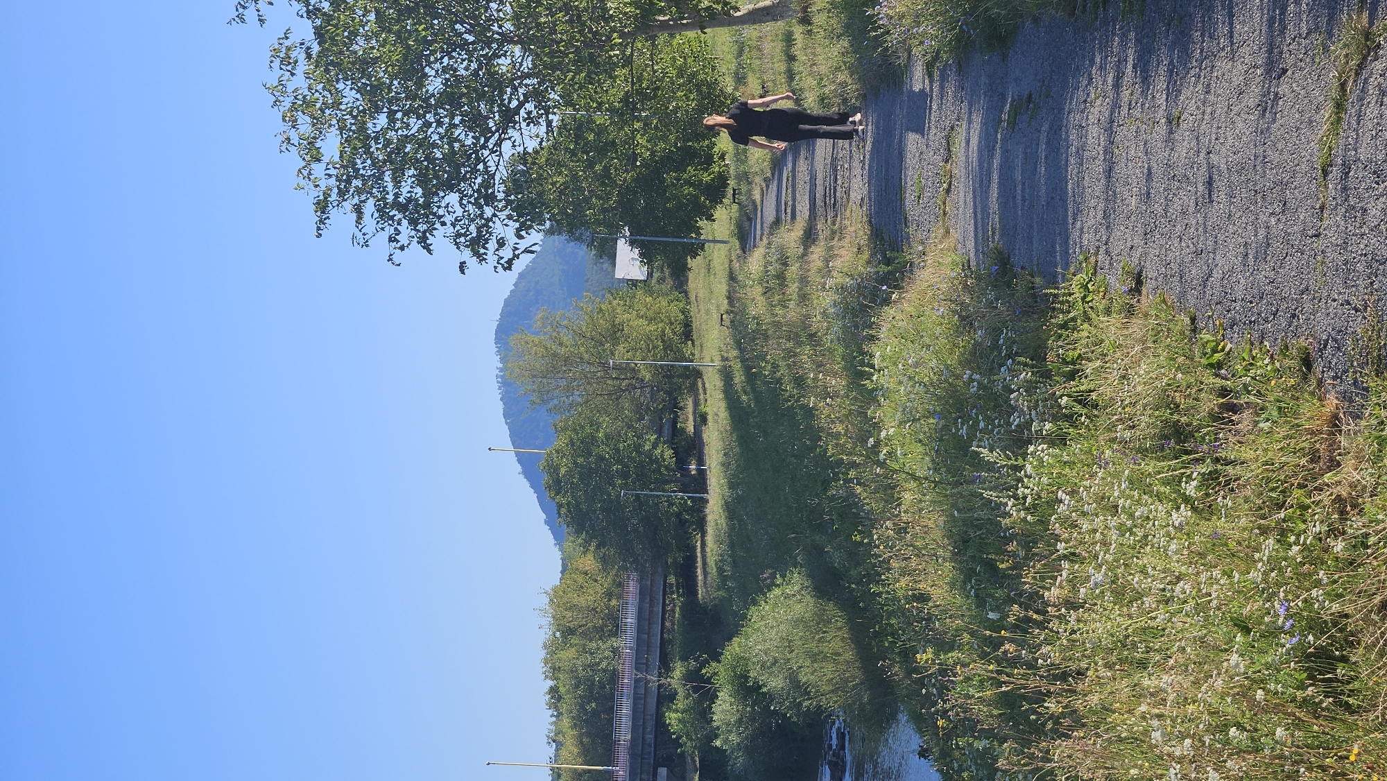 The image depicts a scenic view of the river Hornád in Prešov, Slovakia. A woman is walking along a gravel path that borders the river. The path is flanked by lush green grass and wildflowers. In the background, a bridge spans the river, and a hill rises in the distance, partially covered by trees. The sky is clear and blue.