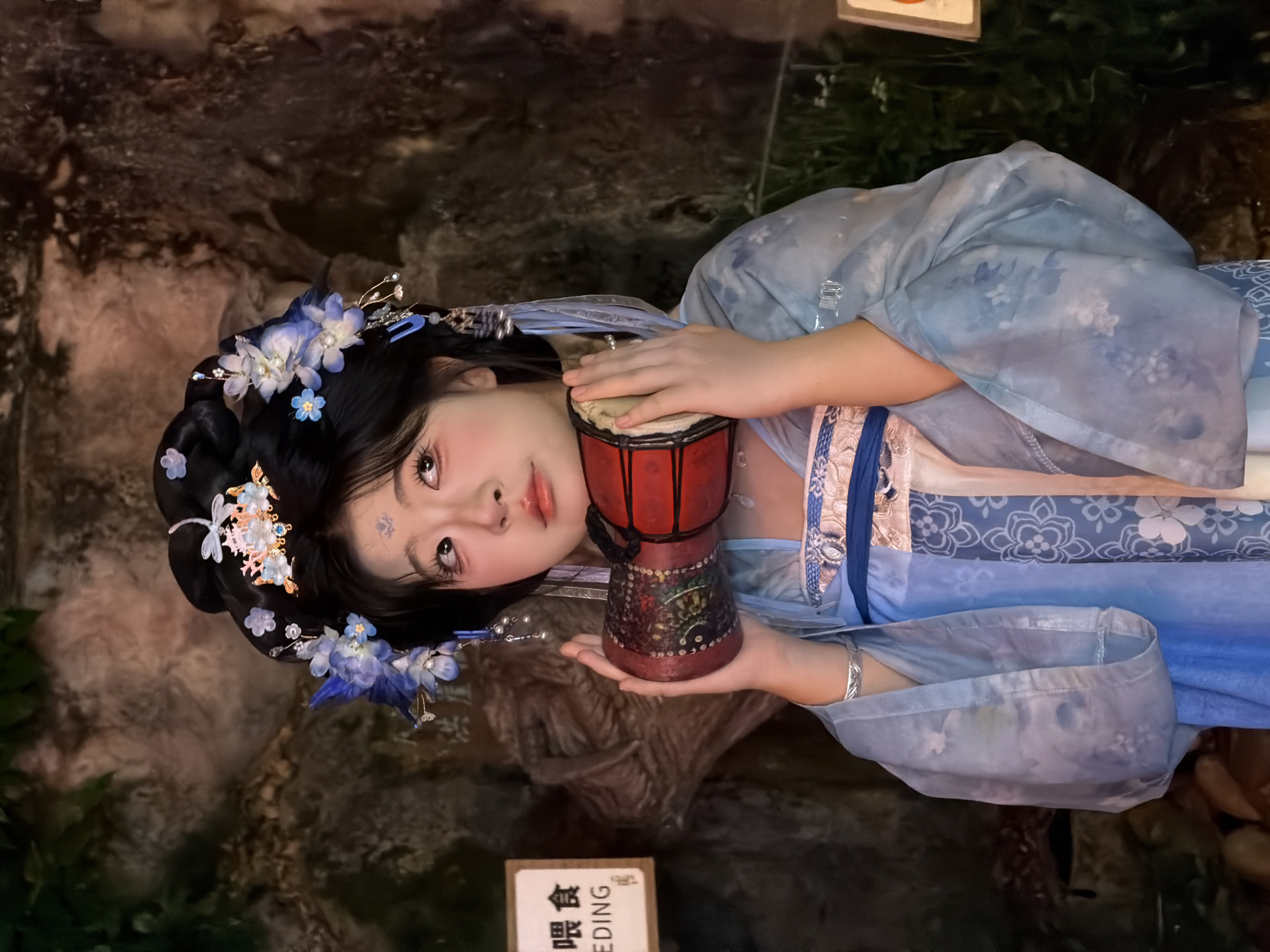 A young East Asian woman, adorned in traditional Chinese attire including an elaborate floral headdress and a light blue garment with intricate patterns, holds a small, red, cylindrical drum. She has fair skin, dark hair styled up, and wears visible makeup with rosy cheeks and dark eyeliner. A silver bracelet adorns her left wrist. The background consists of a rough, rocky wall and lush green foliage, suggesting an outdoor or naturalistic setting. To the left, a sign with Chinese characters and the English word "FEEDING" is visible. The lighting is dim, casting soft shadows and highlighting the woman's face. The image appears to have been taken in the Yuzhong District of China.