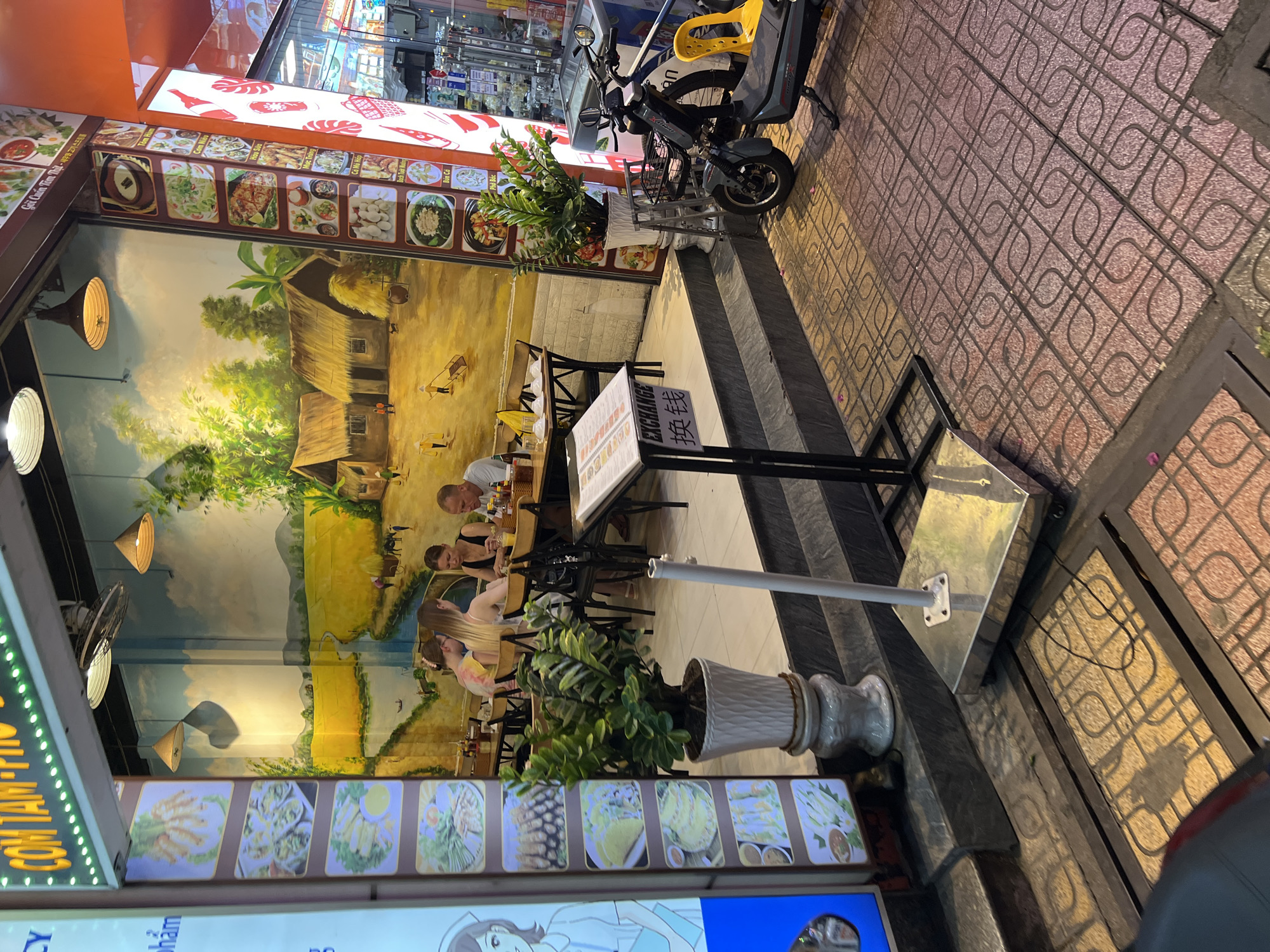 The image depicts a street-level view of a restaurant in Nha Trang, Vietnam. The restaurant features a mural on its interior wall depicting a rural Vietnamese landscape with traditional houses and rice paddies. A menu stand is positioned outside the entrance, displaying food items and also featuring the word "EXCHANGE" in English and Chinese characters. Inside, patrons are seated at tables, engaged in dining. Several potted plants are placed near the entrance, and a motorcycle is parked on the sidewalk. The pavement outside is made of patterned tiles. The overall scene suggests a typical urban setting with commercial activity.