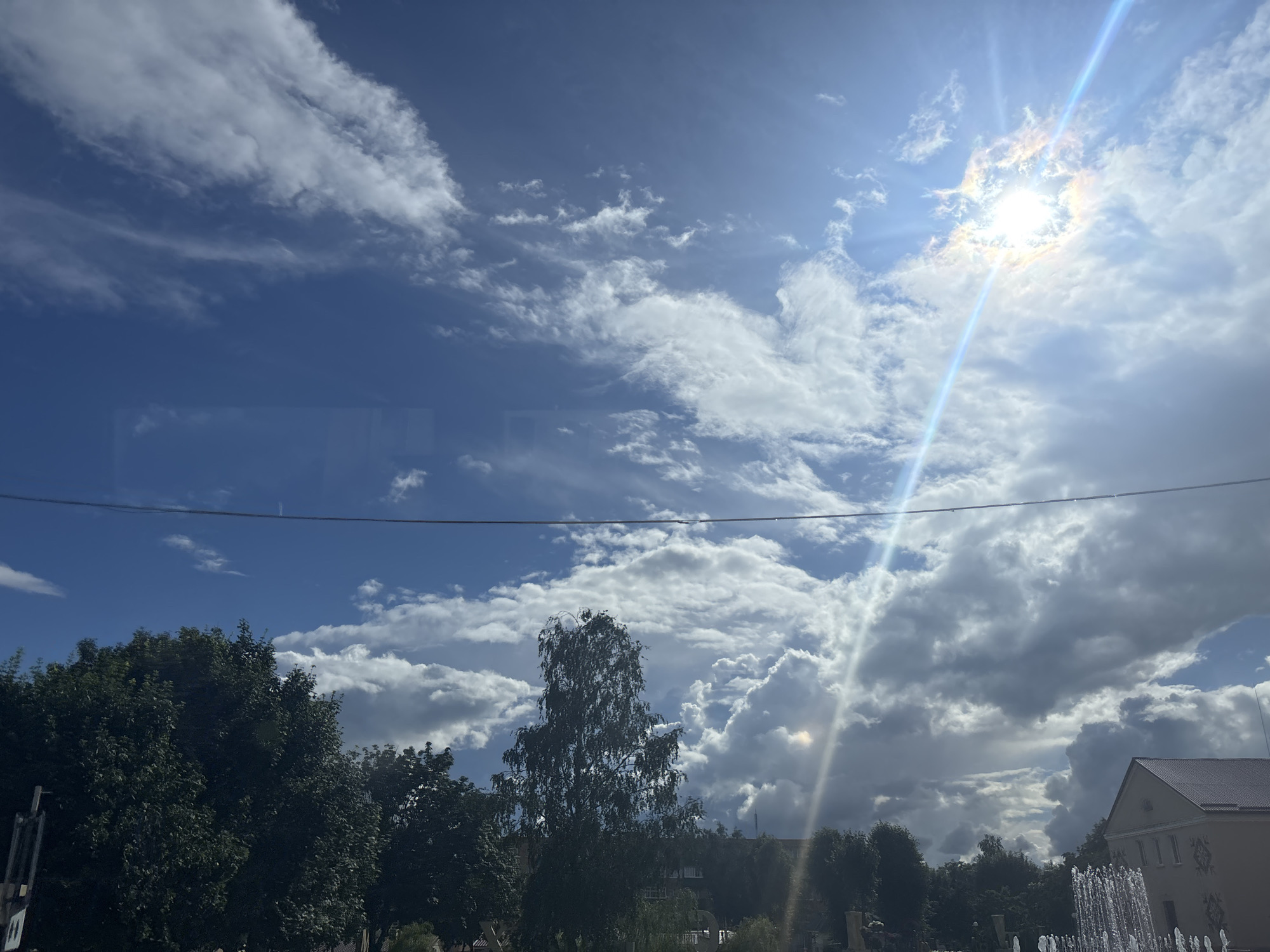 The image captures a view of the sky and cityscape in Kapyĺ, Belarus, on a day with a mix of sun and clouds. The sun is bright, creating lens flares and beams of light that cut across the sky. Fluffy white clouds are interspersed with patches of blue sky, with darker, heavier clouds also visible, suggesting a dynamic weather system.
In the foreground, a line of trees forms a dark silhouette against the brighter sky. Among the trees, a tall, slender tree, possibly a birch due to its pale bark, stands out. In the lower right of the frame, part of a building with a tiled roof is visible, and to its right, a fountain is spraying water, with the droplets catching the sunlight. A thin wire stretches across the upper part of the image. The overall impression is of an outdoor scene in a town or city, likely a public space given the presence of trees and a fountain.