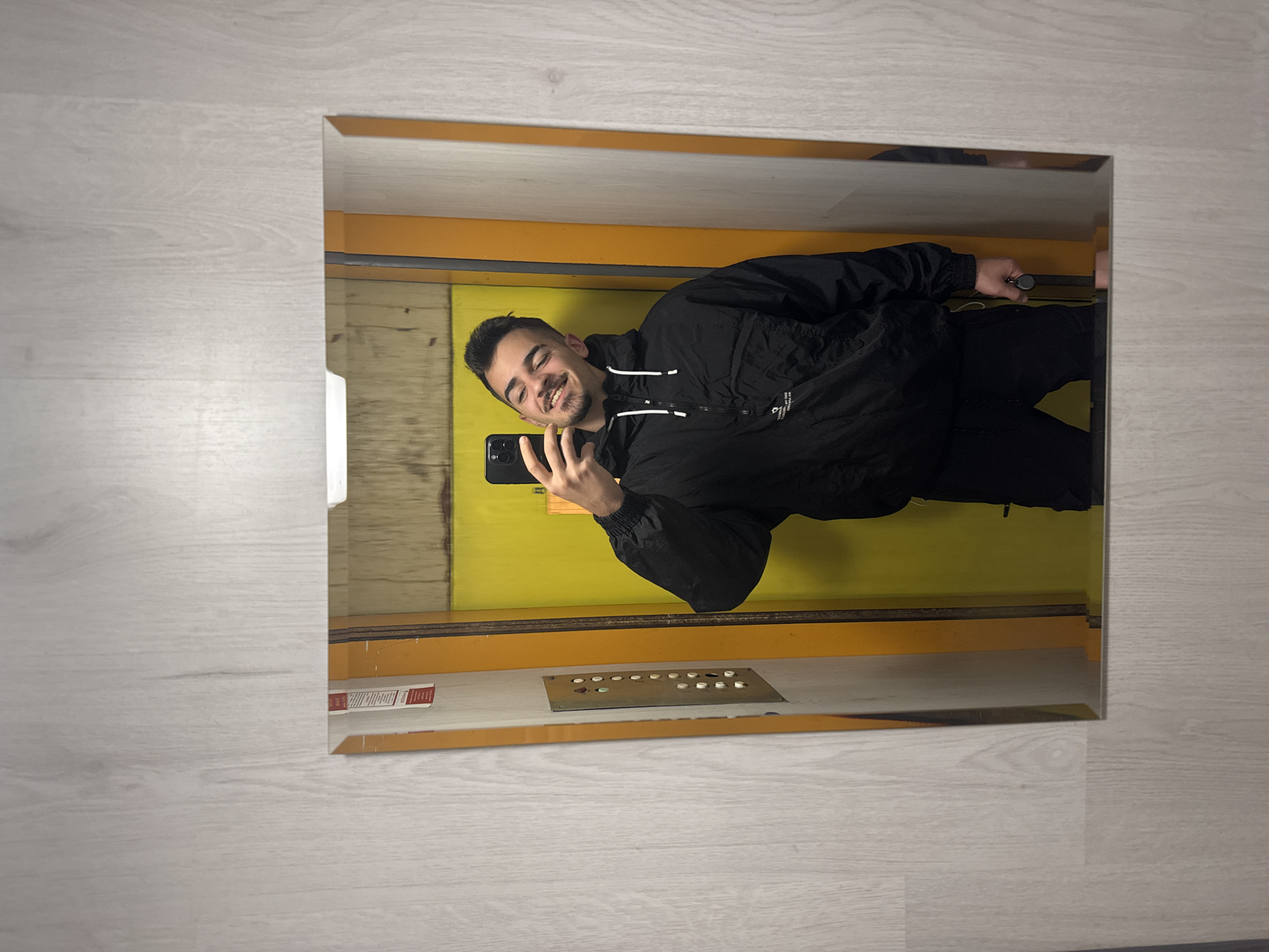 The image captures a young man taking a mirror selfie inside an elevator. He is smiling broadly, holding a black smartphone (an iPhone) with multiple cameras in his left hand to photograph his reflection. The man has short dark hair, a light beard, and is wearing a black hooded jacket, possibly a windbreaker, with some faint white text on the front, and black trousers.
The elevator interior features prominent bright yellow or orange panels on the walls. The selfie is taken in a large, reflective surface, likely a mirror or polished metallic wall within the elevator. Reflected on the lower part of the elevator interior, a control panel with two rows of circular buttons and a larger square button is visible; some buttons appear illuminated, suggesting they are pressed. To the left of the control panel, a small red and white rectangular label or sticker is present, but its text is illegible. The reflection of the man's right hand is visible holding a black keychain or small object near the elevator door frame.
The image is oriented sideways, with the elevator interior appearing horizontally. The area surrounding the elevator's reflective surface, from which the photo was presumably taken, shows light-colored wooden flooring. While the image content does not offer specific visual cues, the location context provided states it is Sofia, Bulgaria.