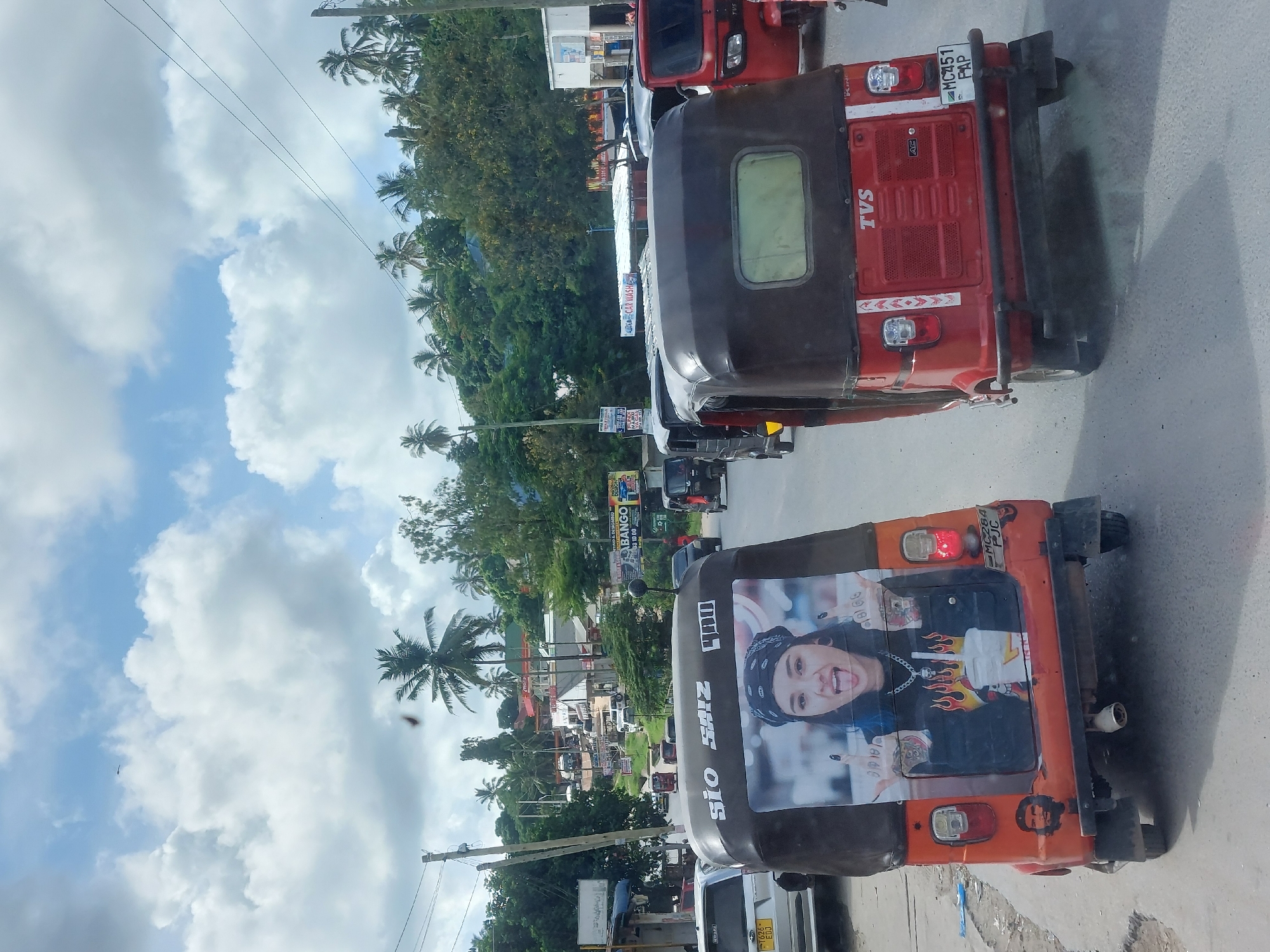 The image captures a vibrant street scene in Dar es Salaam, Tanzania. The focal point is a line of three-wheeled vehicles, commonly known as "Bajajs" or "Tuk-Tuks." The Bajaj in the foreground features a large advertisement on its rear, showcasing a picture of SIO SAIZ. The Bajaj behind it is red and bears the TVS logo. Both have license plates visible. Street scenes are visible, with palm trees lining the roads and various buildings, including shops with signs like "BANGO" visible. The sky overhead is partly cloudy.