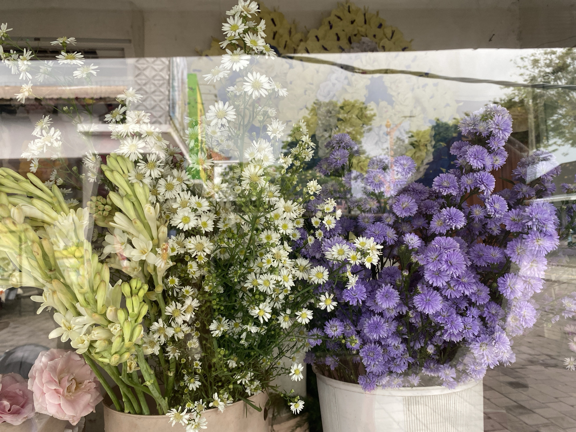 The image captures a vibrant display of fresh cut flowers viewed through a reflective glass window, likely in a shop or market in Kuta Selatan, Indonesia. The foreground is filled with large bouquets arranged in pots.
To the left, there are abundant stalks of white to pale green, tubular-shaped flowers, some beginning to open, reminiscent of tuberoses. Below these, a soft pink rose bloom is partially visible. Centrally, a dense bunch of small white daisy-like flowers with yellow centers stands out, contributing to a fresh and bright aesthetic. To the right, an equally large cluster of small purple daisy-like flowers, possibly asters, adds a contrasting cool tone.
The glass surface creates prominent reflections, layering the outdoor environment over the flower display. Visible reflections include parts of a building structure with a window frame, what appears to be a yellow corrugated roof or awning in the upper background, and the textures of a paved area or sidewalk. Patches of green foliage and trees are also reflected, suggesting an outdoor setting beyond the window. A strong horizontal line, possibly a beam or wire, cuts across the mid-section of the reflected scene.
The interplay of the flowers inside and the reflections from outside creates a dynamic and slightly ethereal composition. The lighting suggests it is daytime. There is no visible text in the image.