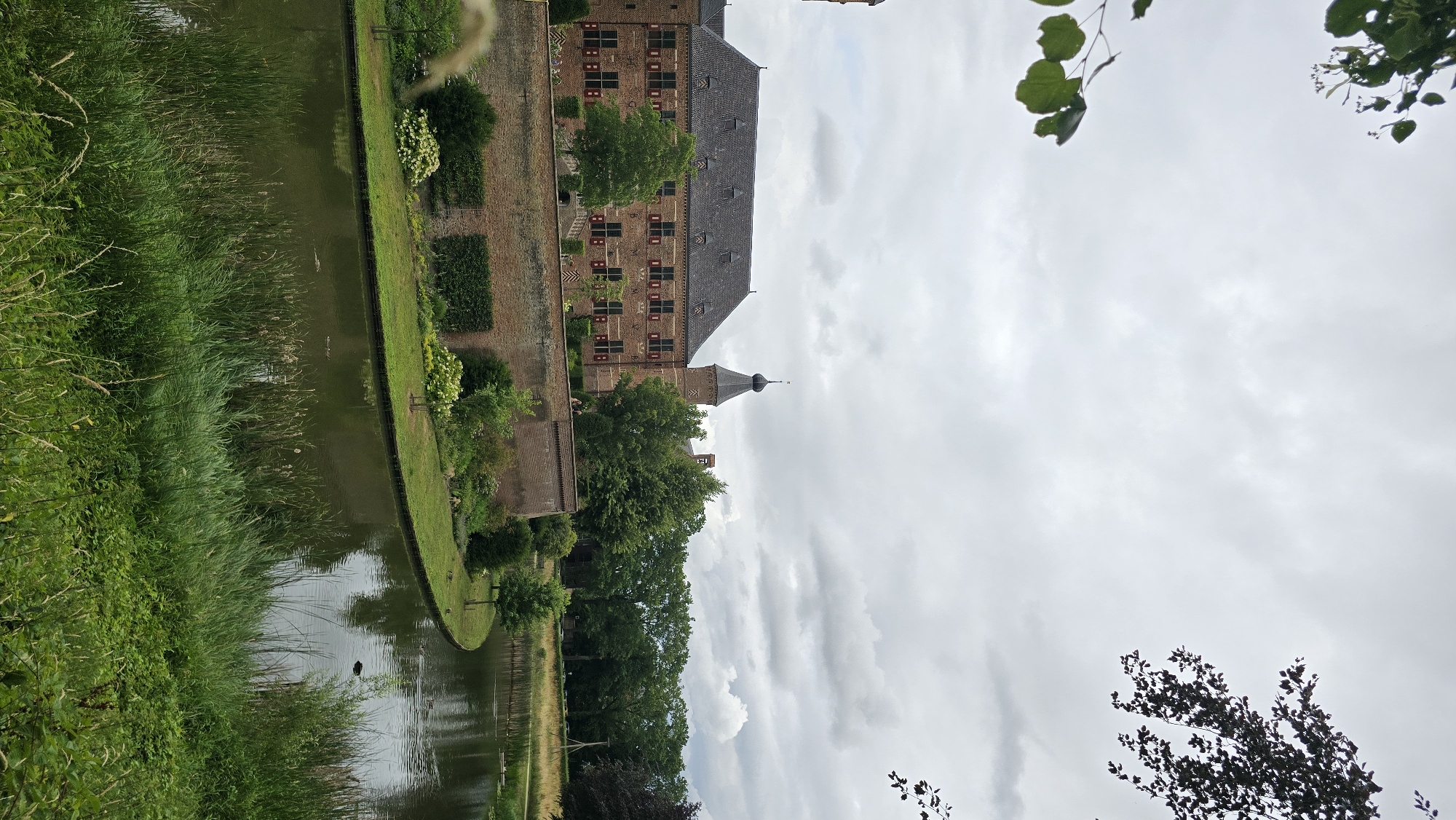 The image depicts a historic brick castle with a dark conical tower, situated in 's-Heerenberg, Netherlands. The castle is surrounded by a moat and a brick wall. Lush green trees and manicured bushes surround the castle, with tall reeds and grasses growing along the edge of the moat. The sky is overcast with clouds. Two figures are visible walking on the castle grounds near the moat. Several ducks are swimming in the moat. The image is taken from a vantage point slightly elevated, looking towards the castle and the water.