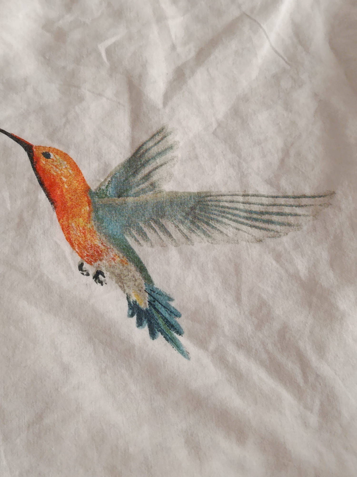 A watercolor painting of a hummingbird is depicted on a white fabric background. The hummingbird is shown in mid-flight with its wings outstretched. Its body is primarily orange and yellow, with accents of blue and green on its wings and tail. The fabric exhibits wrinkles and creases, suggesting it is likely a piece of clothing or upholstery. The image does not contain any information pertaining to the city of Svietlahorsk, Belarus.