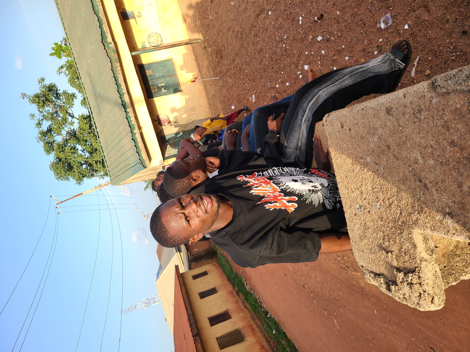A medium-angle outdoor shot captures a group of individuals sitting on a low, rough concrete wall under a clear blue sky, in an unpaved area of Unknown, Unknown. The central figure, a dark-skinned male with short hair, wears a black t-shirt with a red and white "Hellfire" graphic featuring a skull. He is looking directly at the camera with an intense expression, his mouth slightly open, wearing dark pants with a lighter wash pattern and black sandals. His right hand rests on the concrete surface.
Behind him, several other individuals, also seated on the concrete, are partially visible. One person immediately behind him is seen in profile, looking left. Further down the line, a child in a yellow t-shirt and red shorts stands on the reddish-brown dirt ground, facing away from the camera. The ground is scattered with white debris.
In the background to the left, a single-story, light brown building with a corrugated reddish-brown roof has three dark, rectangular windows. To the right, a yellow-painted building with a green corrugated metal roof features faded murals: a red apple labeled "A HPL" and a blue cup next to a soccer ball labeled "BALL." This building has two dark green louvered windows. A thin wooden pole with a small red flag is planted in the dirt near the yellow building.
Overhead, multiple black power lines stretch across the blue sky, connected to a wooden utility pole on the right. A white lattice tower is visible on the distant horizon to the left. Green trees are present behind the buildings and on the horizon. The scene is illuminated by strong sunlight, casting defined shadows.