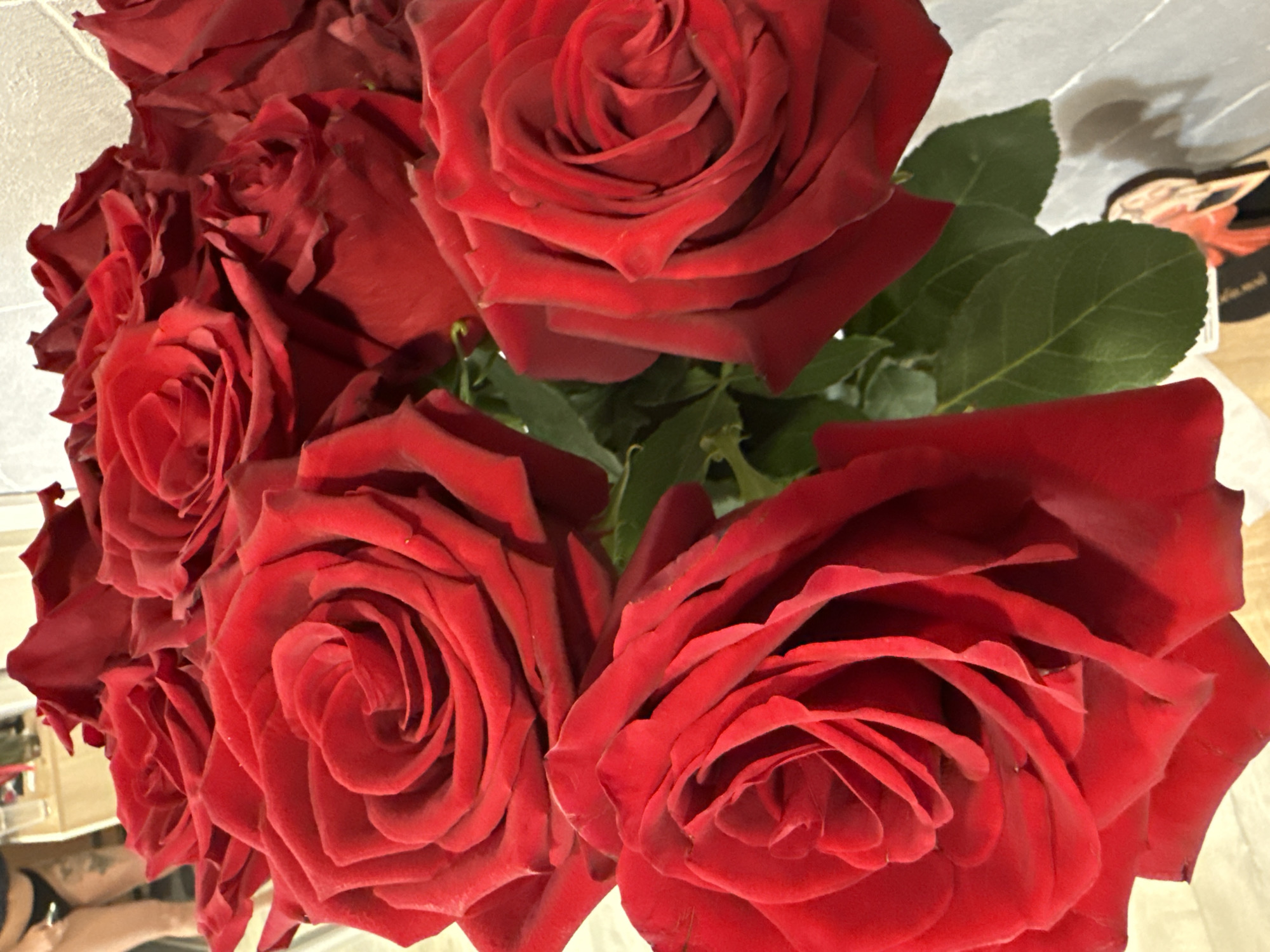 The image shows a close-up of a bouquet of deep red roses with visible green leaves. The roses are in full bloom. The background is out of focus. There is no other indication of the location being Chaikovsky, Russia. There are no people visible in the primary focus of the image.