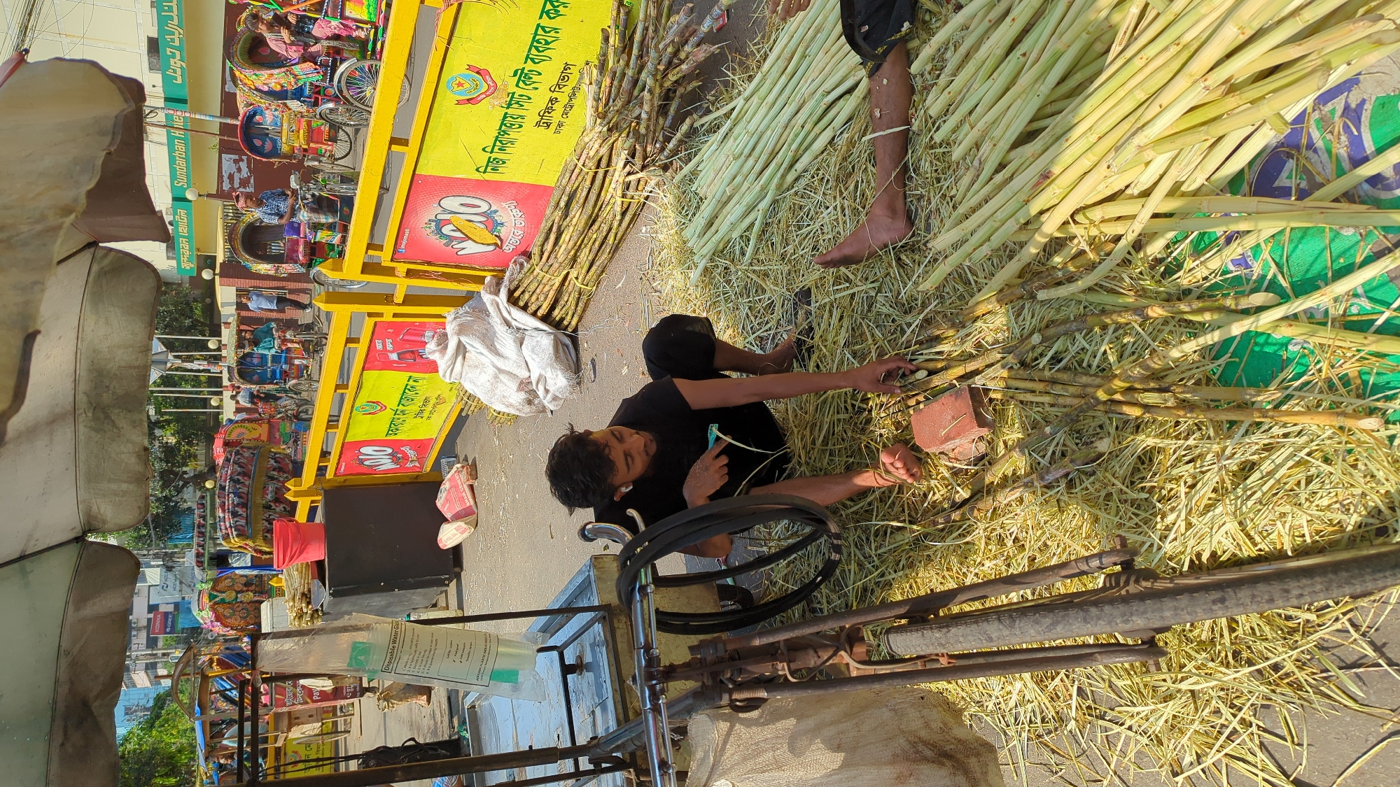 In Dhaka, Bangladesh, a man in a black t-shirt and pants sits on a straw-strewn street, barefoot, cutting sugar cane stalks with a small knife. He is surrounded by piles of cut sugar cane. Next to him, there is a three-wheeled cart with a rusty metal frame and a plastic water container, possibly his workstation. In the background, there are colorful cycle rickshaws parked along the street. The area appears to be a bustling commercial district with signs in Bengali. The background includes signs with the text "Sundarban Hotel" and "নিজ নিরাপত্তায় সিট বেল্ট ব্যবহার করু" which translates to "Use seat belts for your safety". Also included is the phrase "অকারণে হর্ন বাজাবেন না" which means "Do not honk unnecessarily."