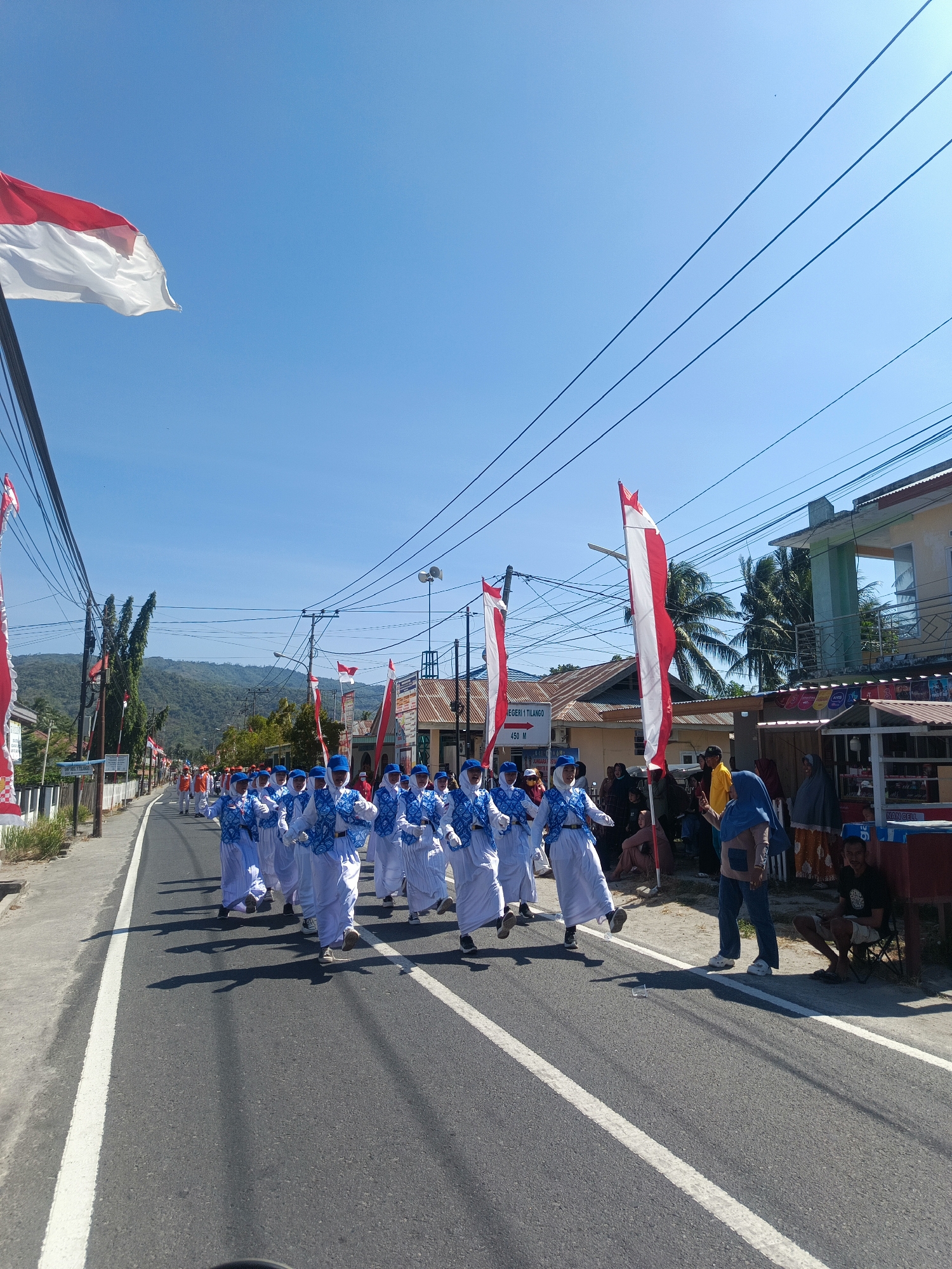 A parade is taking place on a paved road in the city of Tenggela, Indonesia. The sky is clear and blue, with a few wispy clouds. The road is lined with buildings and palm trees. The parade features a group of people marching in unison, dressed in white and blue uniforms with red and white flags displayed along the route. Several spectators are watching the parade from the side of the road.