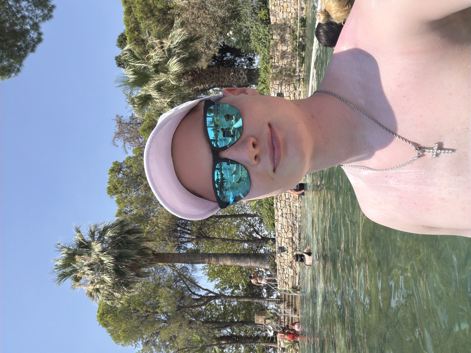 In Pamukkale, Turkey, a young person wearing a white cap and sunglasses is taking a selfie in a clear, shallow body of water. The water reflects a blue sky, palm trees, and other swimmers. The person is wearing a silver cross necklace. In the background, there are stone structures, stairs leading into the water, and lush green trees. Several other people are visible in the water and on the stairs, some swimming and others standing. The sun is shining brightly, creating a warm and inviting atmosphere.