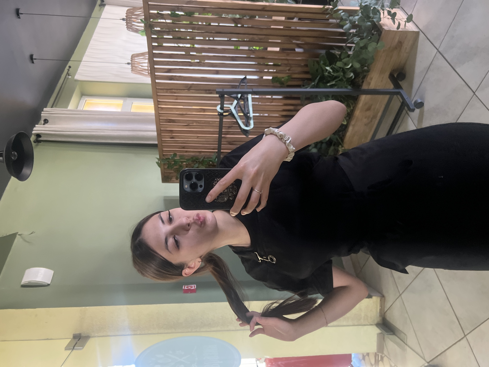 A young adult female is depicted in a mirror selfie taken indoors in Красные Баки, Russia. She has dark brown hair pulled into a ponytail, which she holds with her left hand. Her face is turned towards the camera, and she is making a puckered lip expression. She wears a black short-sleeved top or dress featuring a distinctive gold-colored metal accessory on the left chest. A thin gold ring is on her right ring finger, and a light-colored, ruffled fabric bracelet, along with a thin gold bangle, adorns her left wrist. She holds a black iPhone, likely a Pro model, in her right hand, which has a case featuring an embossed eagle or crest design.
The background, visible through the mirror, reveals an interior space with pale green walls and a dark grey ceiling. On the left, a pale yellow wall is visible, along with part of a glass door or panel featuring a circular design and faint, reversed Cyrillic text "ЕВРАЗИЯ". A red fire alarm box is mounted on the green wall. Various lighting fixtures are present: two black industrial-style pendant lamps hang from the ceiling, and two lower pendant lamps with light-colored, possibly wicker or wooden, shades are situated over a wooden slatted room divider. A window with light-colored curtains is visible, allowing bright light to enter. Green foliage is arranged in wooden planter boxes along the base of the divider. A black metal clothing rack on wheels stands behind the woman, holding two hangers (one light blue/green, one dark). The floor is covered with light-colored square tiles.