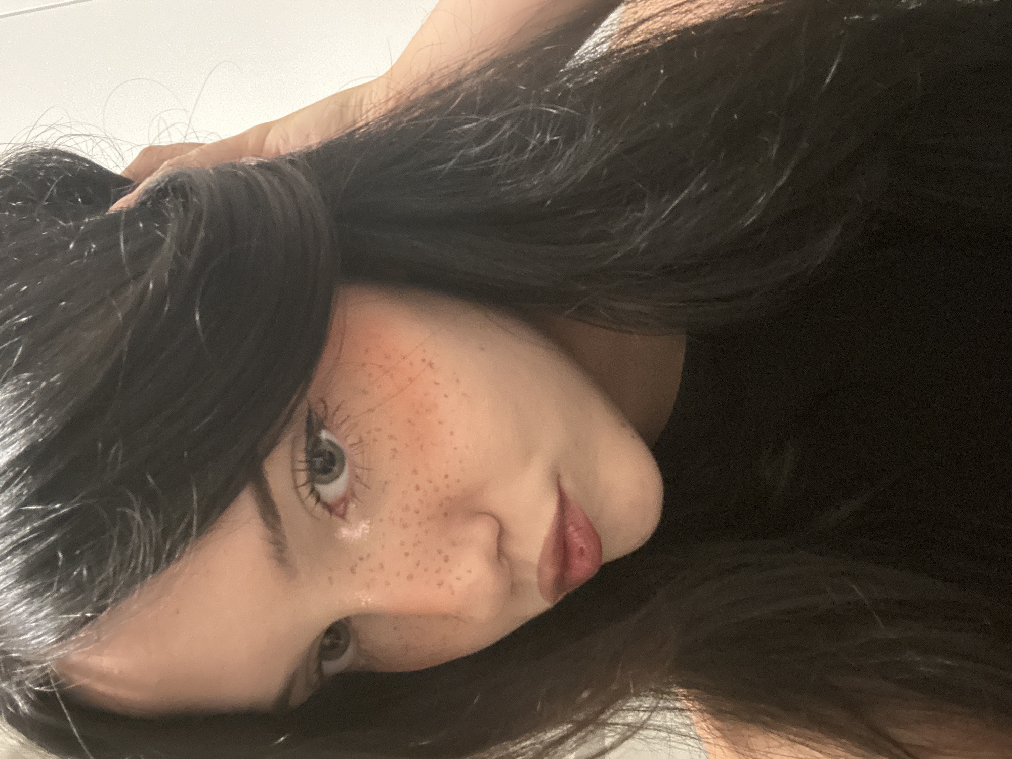 A young woman with long, dark hair and grey eyes is shown in a close-up portrait. She has freckles across her nose and cheeks and is wearing subtle makeup, including eyeliner and blush. Her lips are slightly parted, and she is looking off to the side. The background is a plain white wall. There is no indication of the city of Cupra Marittima, Italy, in the image.