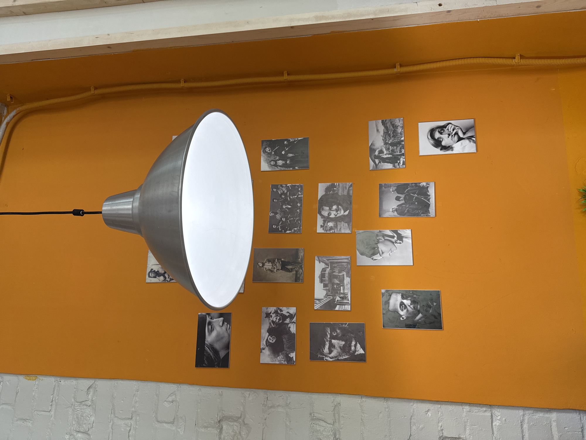 A well-lit, medium shot captures a portion of an interior space. An orange wall is adorned with various black and white photographs, possibly portraits, arranged in a somewhat random, informal manner. To the left, a large, industrial-style lamp is positioned, its silver-toned shade partially obscuring the images on the wall. The lamp is suspended from a black cord. Above the orange wall, there are visible white beams, and the bottom part of the wall transitions to a white brick texture. A yellow conduit runs along the top of the orange wall. The scene is devoid of people. The location is Moscow, Russia.