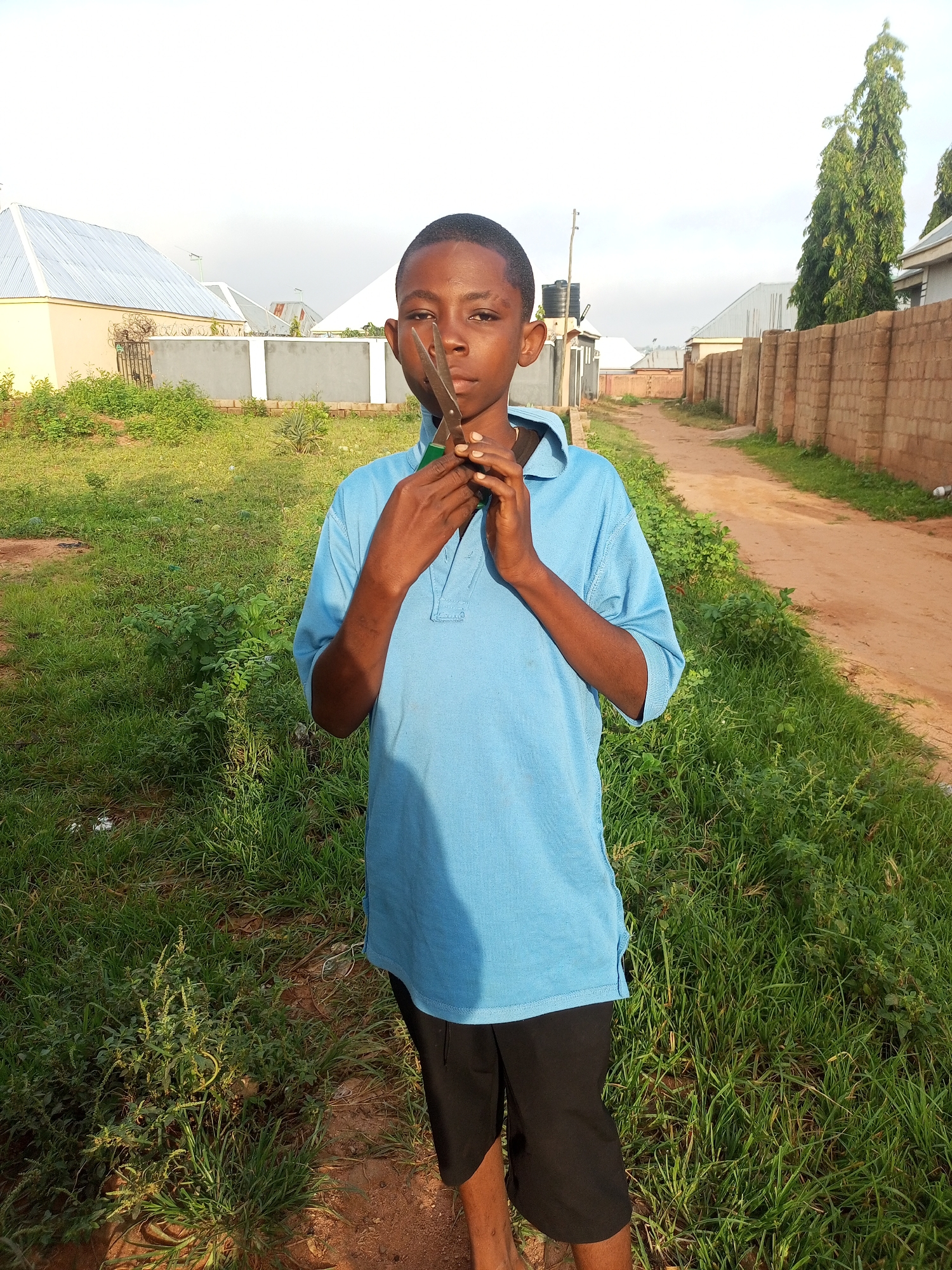 A young boy stands outside in what appears to be a dusty, semi-urban area in Tempure, Nigeria. He is wearing a light blue polo shirt and black shorts. In his hands, he holds a pair of scissors, positioning them near his face. The background shows a dirt road lined with a brick wall on the right and various buildings and structures, including what looks like a water tank, on the left. Lush green grass and some sparse vegetation grow along the sides of the road and in front of the buildings. The sky is overcast, suggesting it might be late afternoon or early morning.