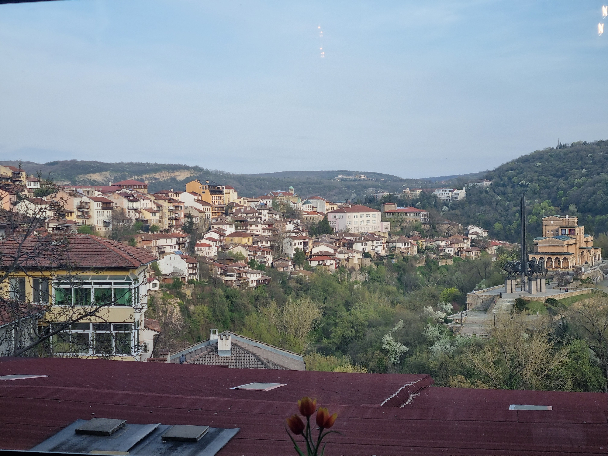The image shows a view of Veliko Tarnovo, Bulgaria, likely taken from a window. A dense cluster of buildings with red-tiled roofs sits on a hill. Lush green trees and vegetation cover the hillside. In the distance, rolling hills are visible under a pale blue sky. A large monument complex with a bronze sculpture of mounted riders and a building with arched openings stands on a nearby hill. Foreground shows the edge of a red roof with windows. Three orange flowers are positioned on the edge of the roof.