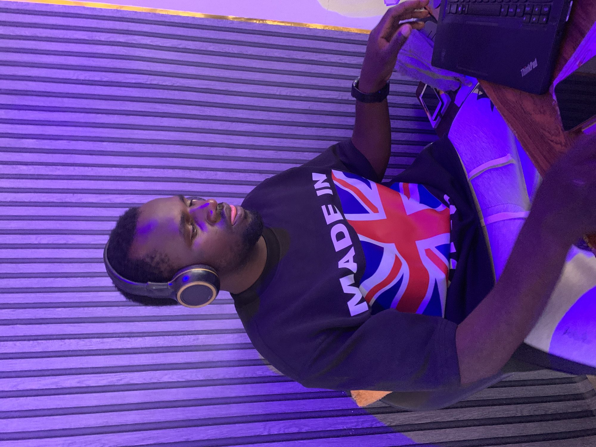 A medium shot depicts a man in an indoor setting, likely in Jos, Nigeria, working on a "ThinkPad" laptop. He is wearing headphones and a dark T-shirt with "Made in" written above a partial British flag. He appears to be seated at a wooden desk. The background features a textured, horizontally striped wall with a purple/blue hue, and there may be decorative gold trim near the ceiling.