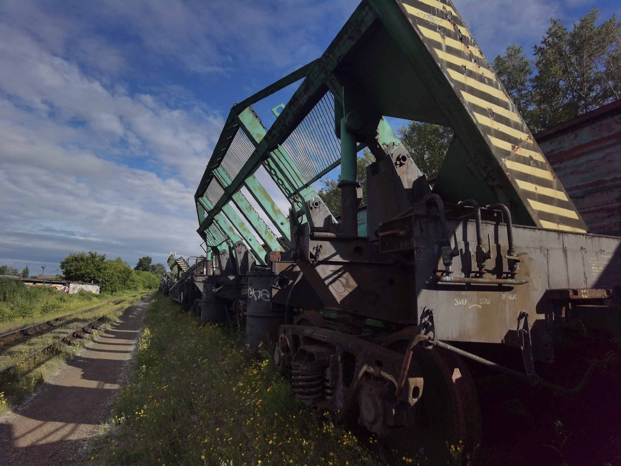 The image depicts a derelict train yard in Chaikovsky, Russia, with several abandoned railway cars. The cars are heavily rusted and overgrown with vegetation, suggesting they have been left undisturbed for a significant period. The sky above is partly cloudy, with patches of blue visible. The train tracks themselves are also in disrepair, with weeds growing between the rails. In the background, some trees and buildings can be seen, hinting at the proximity of an urban or industrial area. The overall impression is one of decay and neglect.