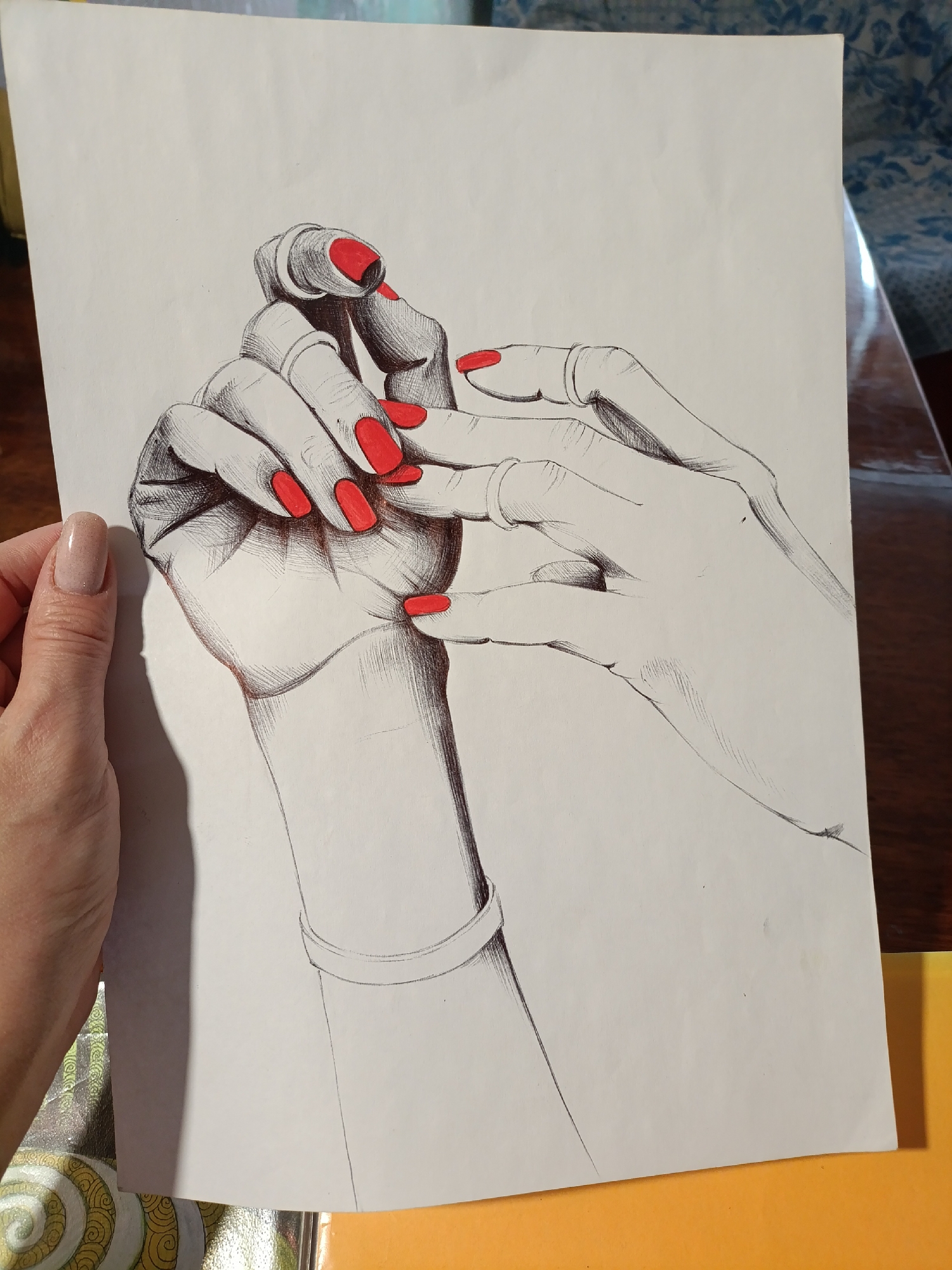 The image displays a drawing of two hands intertwined, rendered in black ink with striking red accents on the fingernails. One hand is positioned as if resting on or holding the other, creating a sense of intimacy or connection. The drawing is done on a sheet of white paper, which is held up to reveal its surface. A person's left hand, with a natural-colored manicure, is visible on the left side of the frame, holding the drawing. The background is blurred, showing hints of furniture and fabric, suggesting an indoor setting. The drawing itself is the primary focus, with the artist's detailed use of line work to create shading and form. There are no discernible landmarks or environmental cues to indicate a specific city or location.