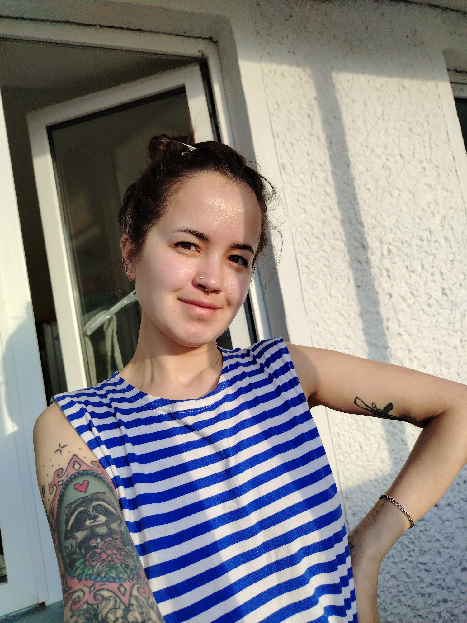 A young woman is captured in a medium close-up outdoor portrait, likely on a balcony or porch, in Noviy, Russia. She has dark hair styled in a bun with a clip and wears a blue and white horizontally striped sleeveless top. Her left arm features a colorful full-sleeve tattoo depicting a raccoon surrounded by flowers within an ornate frame. On her right upper arm, a smaller black tattoo of a stylized rifle or gun is visible, and she wears a silver chain bracelet on her right wrist.
The woman has several ear piercings and two nose piercings: a stud on her nostril and a ring on her septum. She looks towards the viewer with a calm expression and a slight, natural smile. Her face is warmly illuminated by direct sunlight, highlighting her features.
Behind her to the left is a white-framed window or door, slightly ajar. To her right, a textured white stucco wall is visible. The strong, directional sunlight creating distinct shadows on the wall suggests the photo was taken during the golden hour, in the late afternoon or early evening, on a clear, sunny day. No visible text is present in the image.