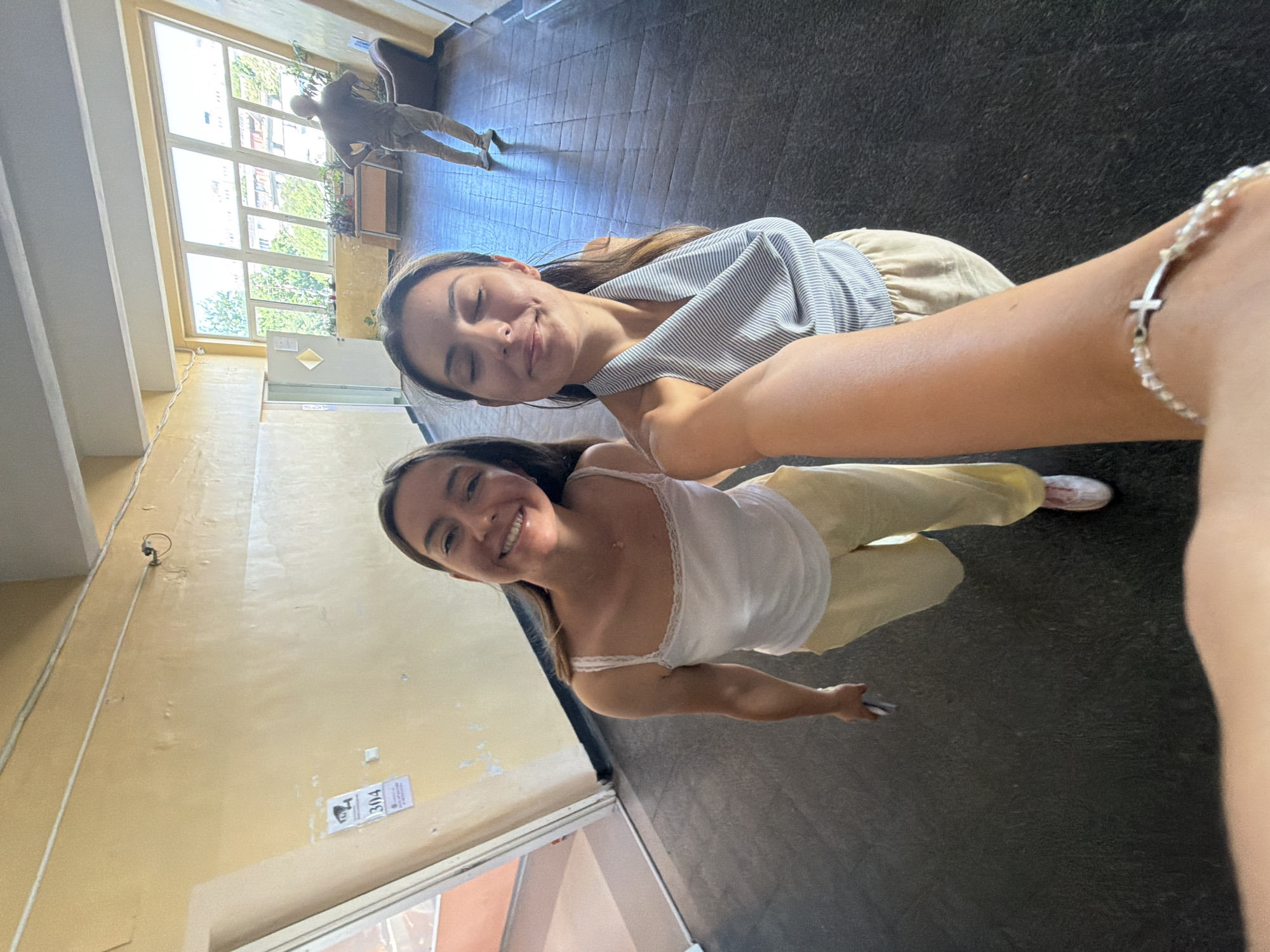 This is a wide-angle, eye-level selfie taken by two young women in a well-lit indoor hallway, likely a school or institutional building. The walls are a pale yellow, with some peeling paint and a sign indicating "304" with a security camera symbol. The floor is dark grey tile. In the background, large, angled windows reveal an outdoor scene with trees and buildings, suggesting an urban setting. A man in a dark shirt and light pants is visible in the distance, standing with his hands on his hips. The woman in the foreground on the right is wearing a gray and white striped halter top and beige pants, her eyes closed and a smile on her face. The woman on the left is wearing a white tank top and yellow pants, looking at the camera with a wide smile. The arm of the person taking the selfie, adorned with a beaded bracelet with a cross charm, is prominent in the foreground. The scene appears to be in Burgas, Bulgaria, based on the provided context, and the style of the building, particularly the windows and hallway design, is consistent with educational institutions found in that region.